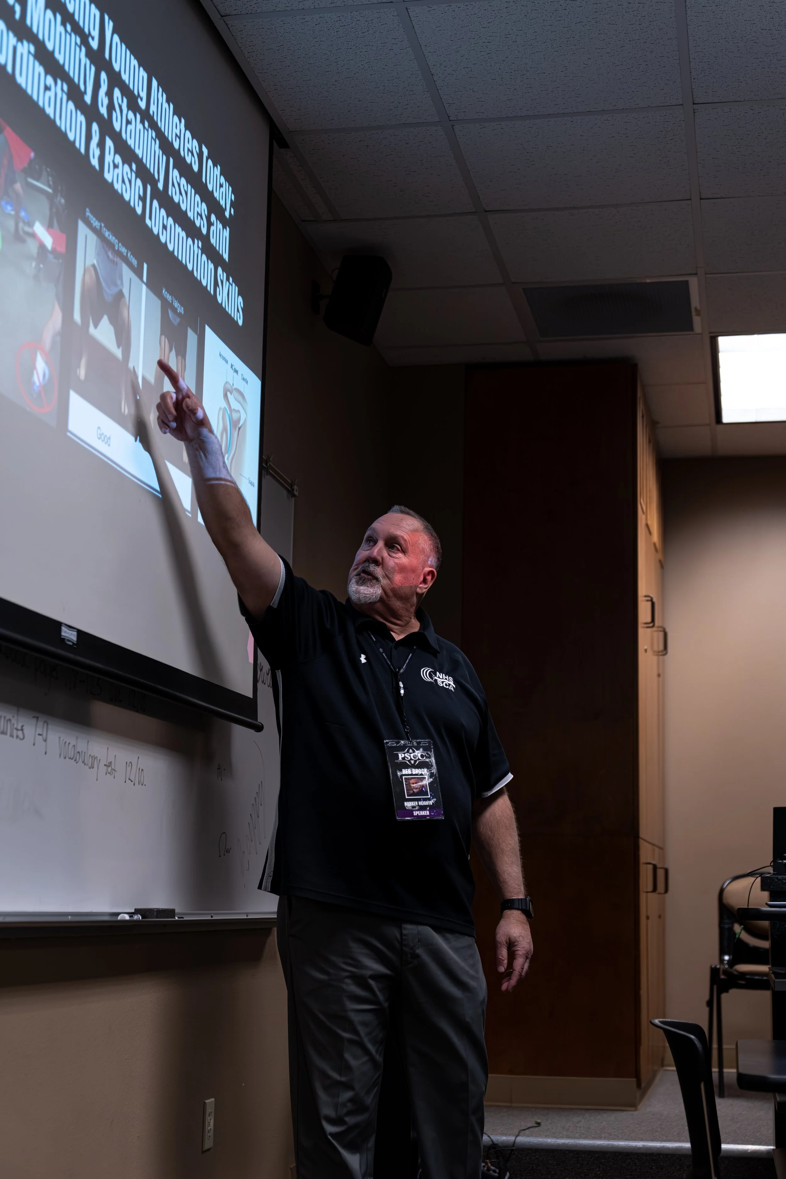A man pointing at a presentation slide on a projector screen in a classroom or conference room.