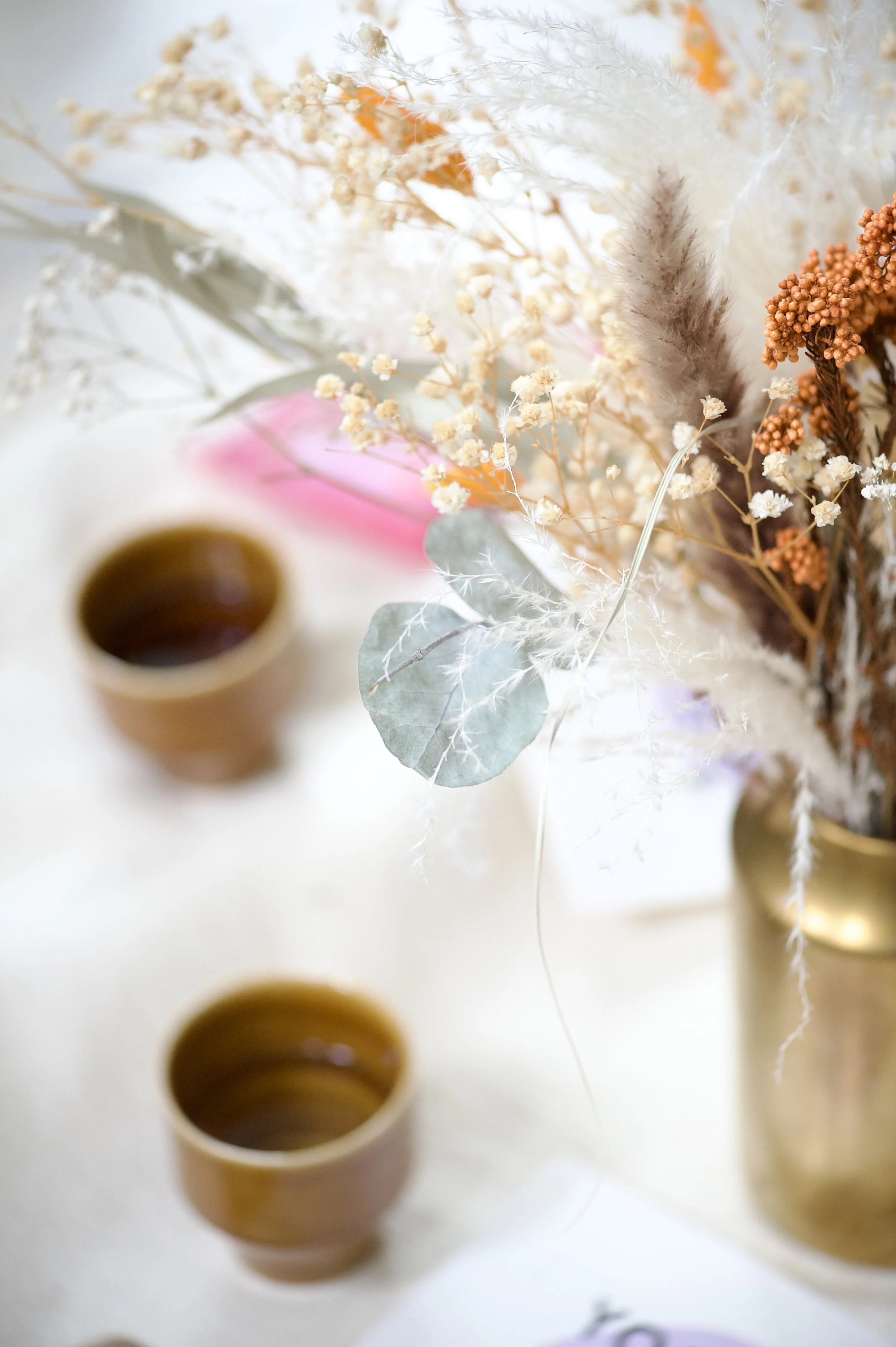 Eine Blumenstrauß in einer goldenen Vase mit getrockneten Blumen und Blättern, neben zwei braunen Tassen mit einem dunklen Getränk auf einem hellen Tisch.