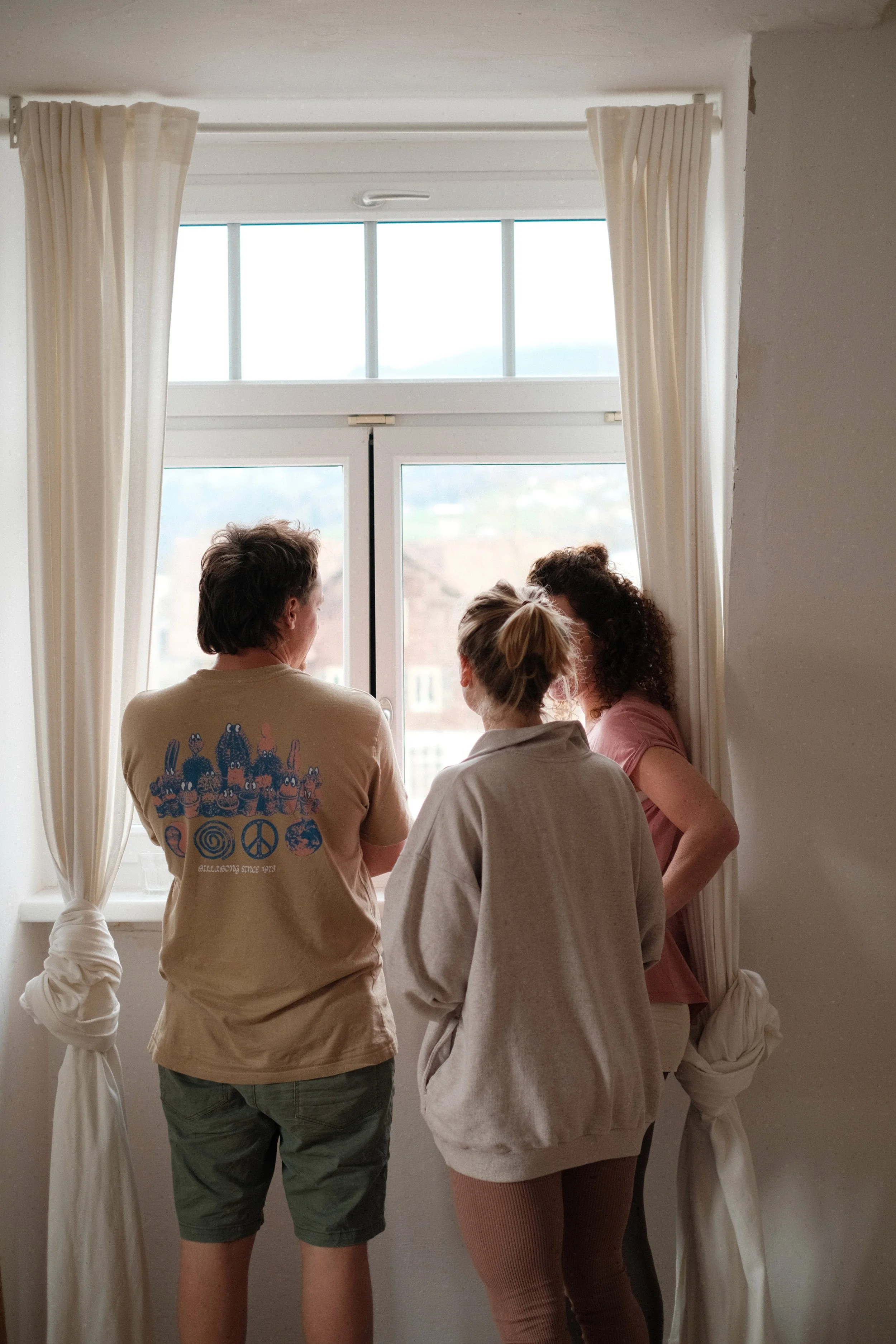 Drei Menschen stehen vor einem Fenster, schauen nach draußen, im Hintergrund eine Stadt und Berge, weiße Vorhänge, hell erleuchteter Raum.