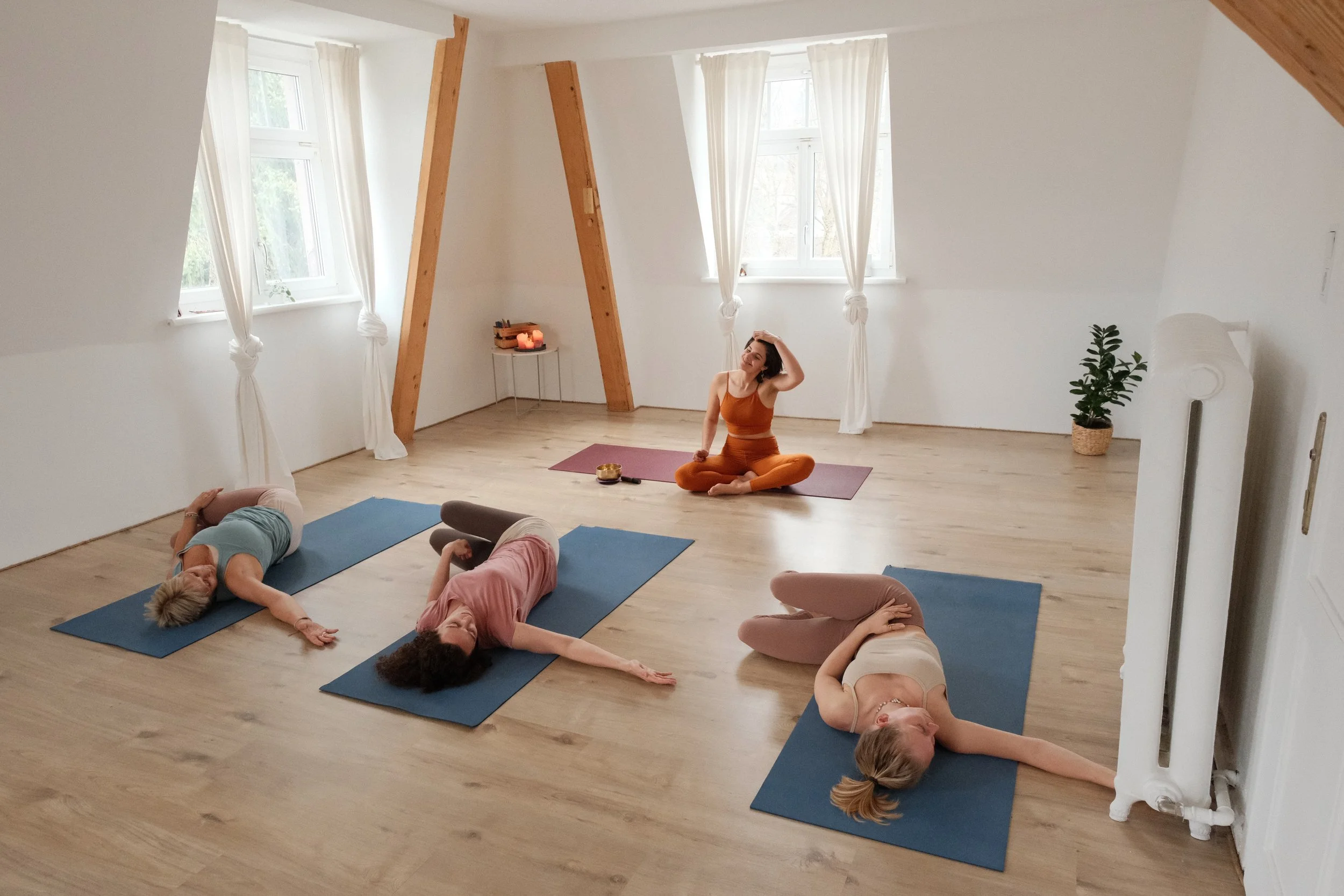 Vier Frauen machen Yoga in einem hellen, minimalistischen Raum mit Holzboden und weißen Wänden, in der Nähe von Fenstern mit weißen Vorhängen.