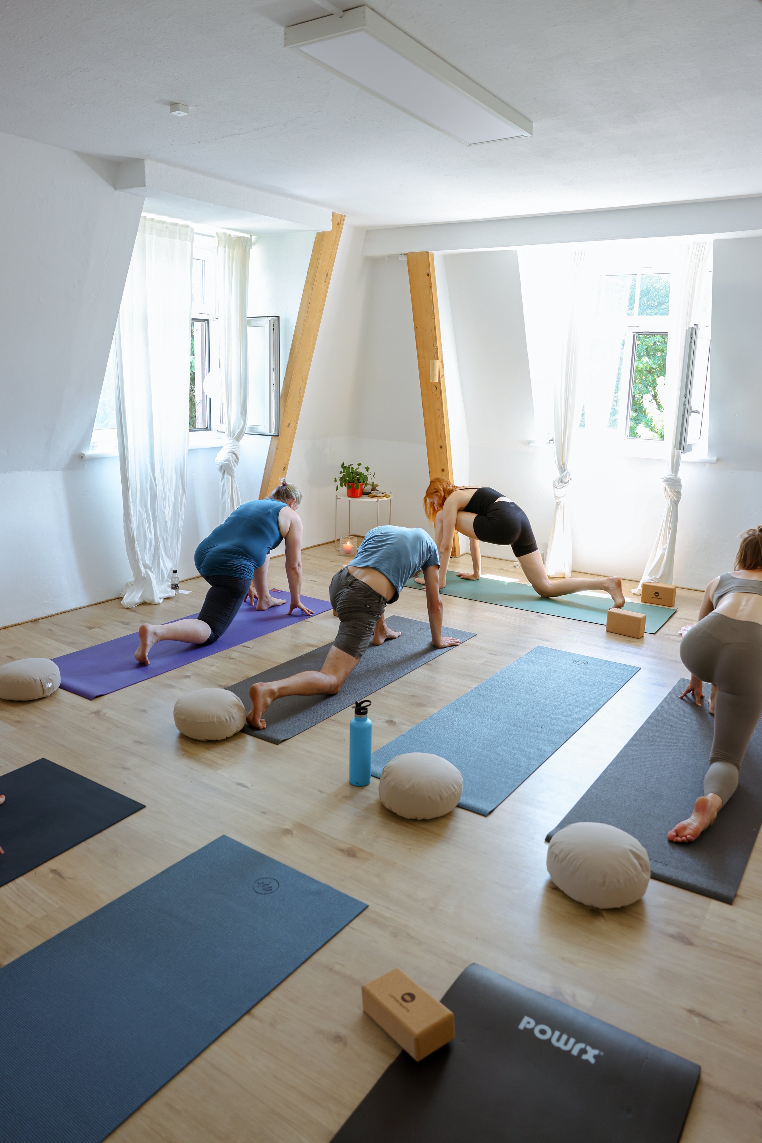 Menschen in einem Yoga-Kurs in einem hellen Raum mit Holzboden und großen Fenstern, die Yoga-Posen einnehmen.