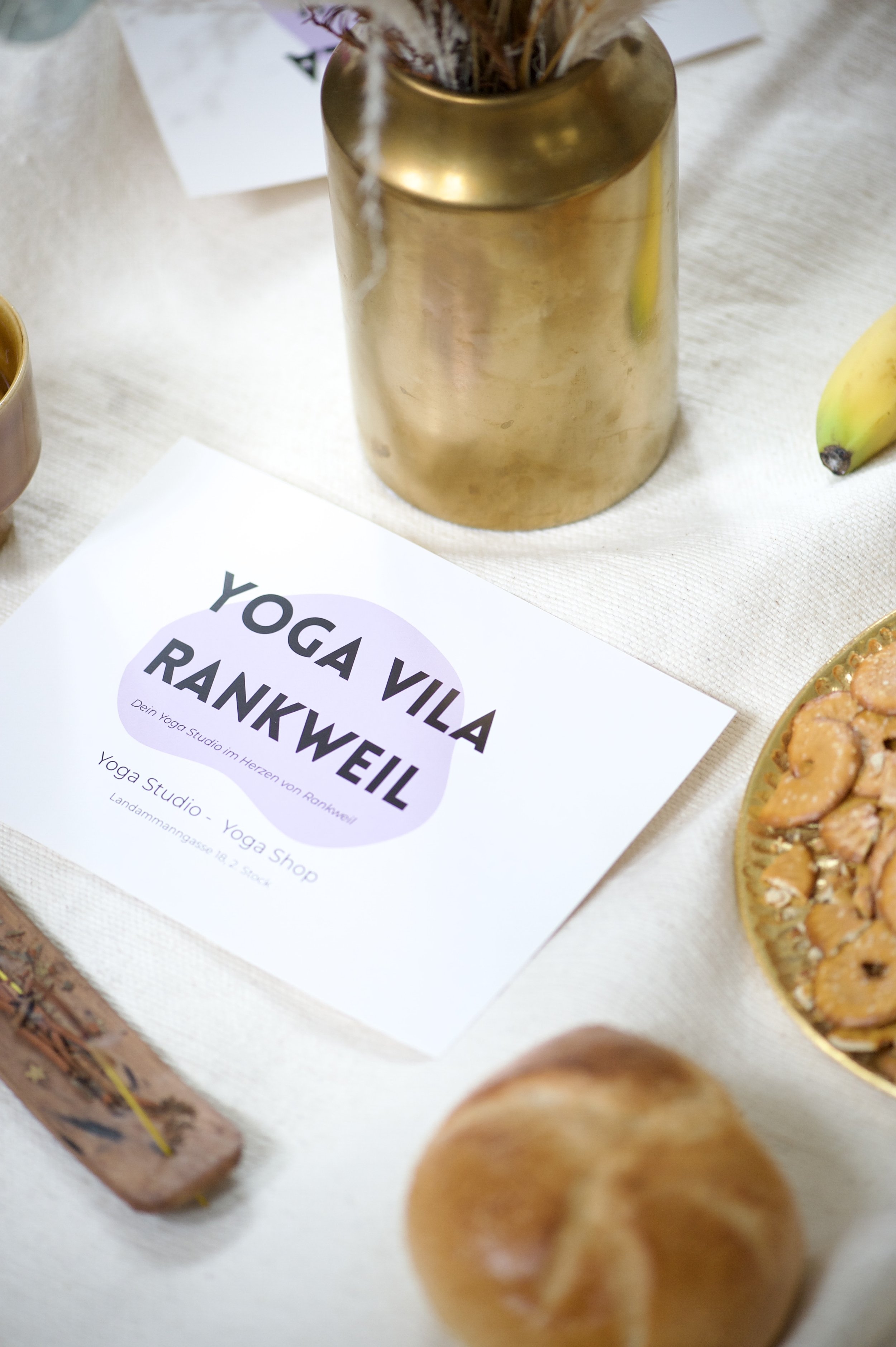 Karte mit der Aufschrift "Yoga Vila Rankweil" auf einem Tisch, umgeben von goldfarbenen Vasen, einem Banane, einem Brötchen und Snackschüssel.
