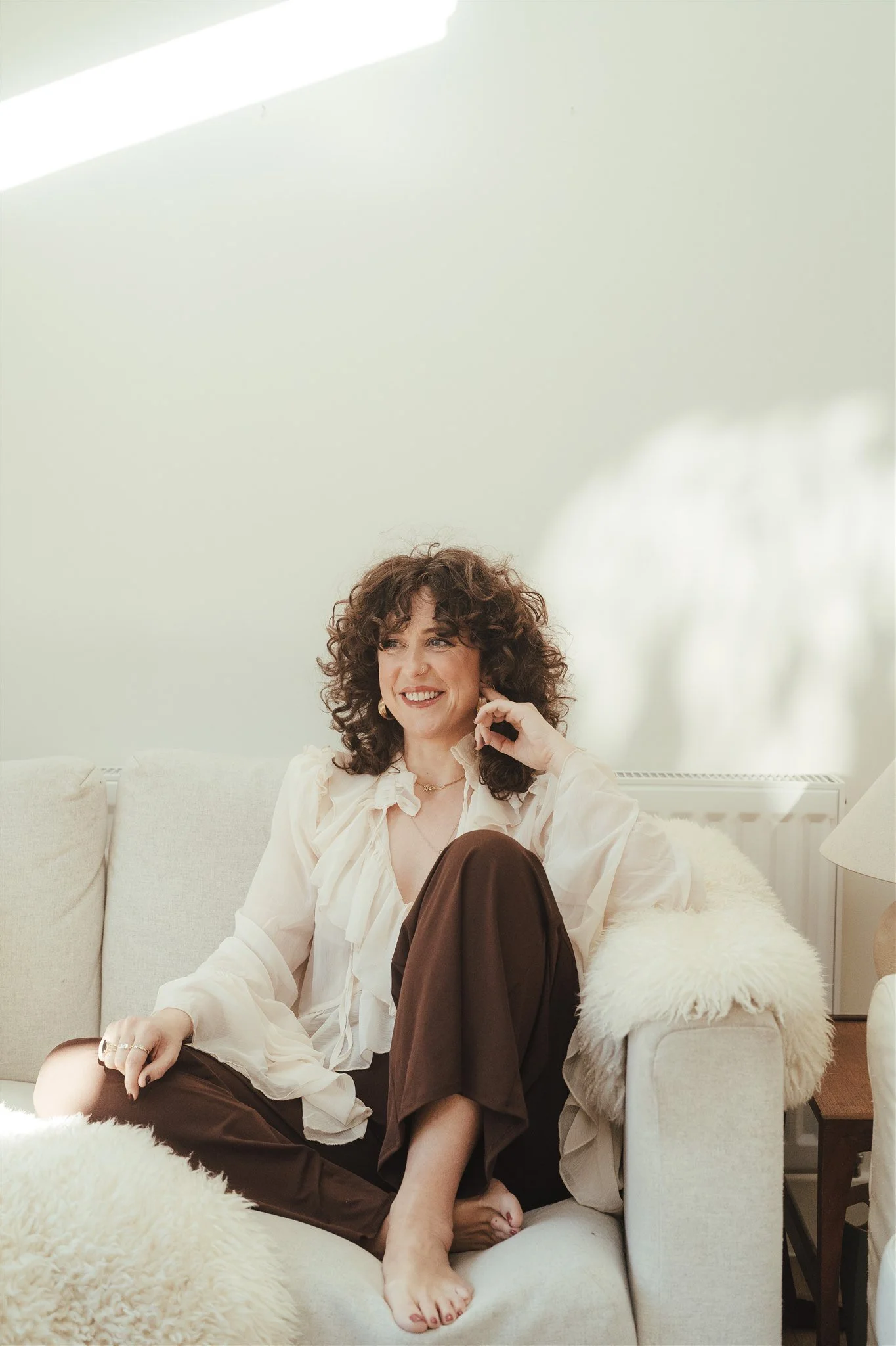 A woman sitting on a cream-colored couch with a fluffy blanket, smiling and looking to her left. She has curly brown hair and is wearing a cream blouse and dark pants.