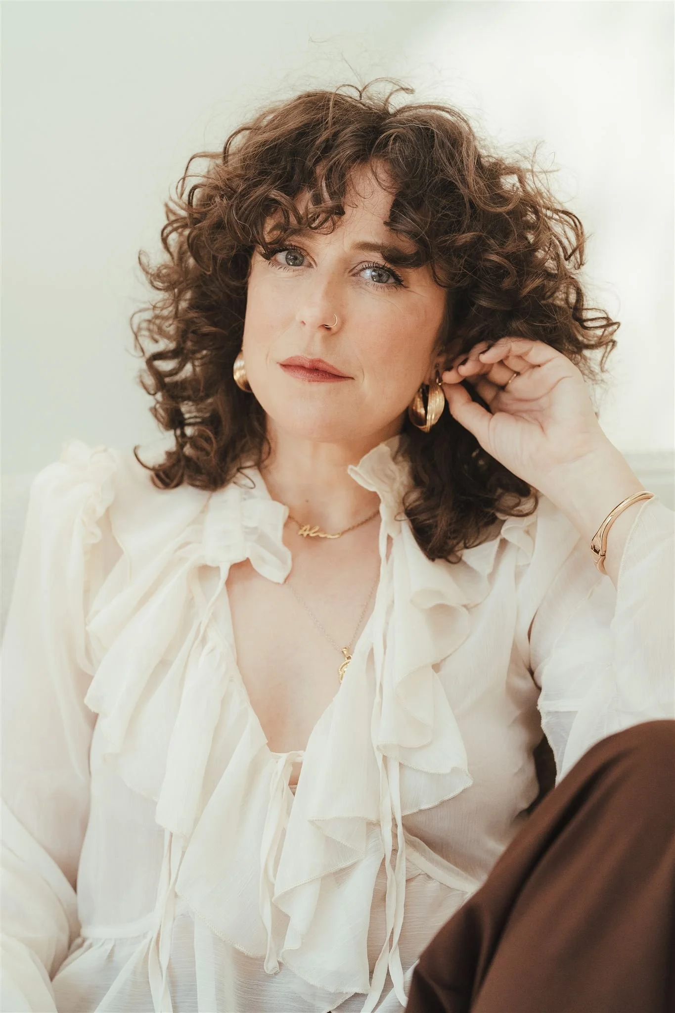 A woman with curly brown hair, blue eyes, and light skin is adjusting her earring. She is wearing a white blouse with ruffles, gold hoop earrings, a gold necklace, and a bracelet. There is a person in a brown shirt partially visible in the lower right corner of the image.