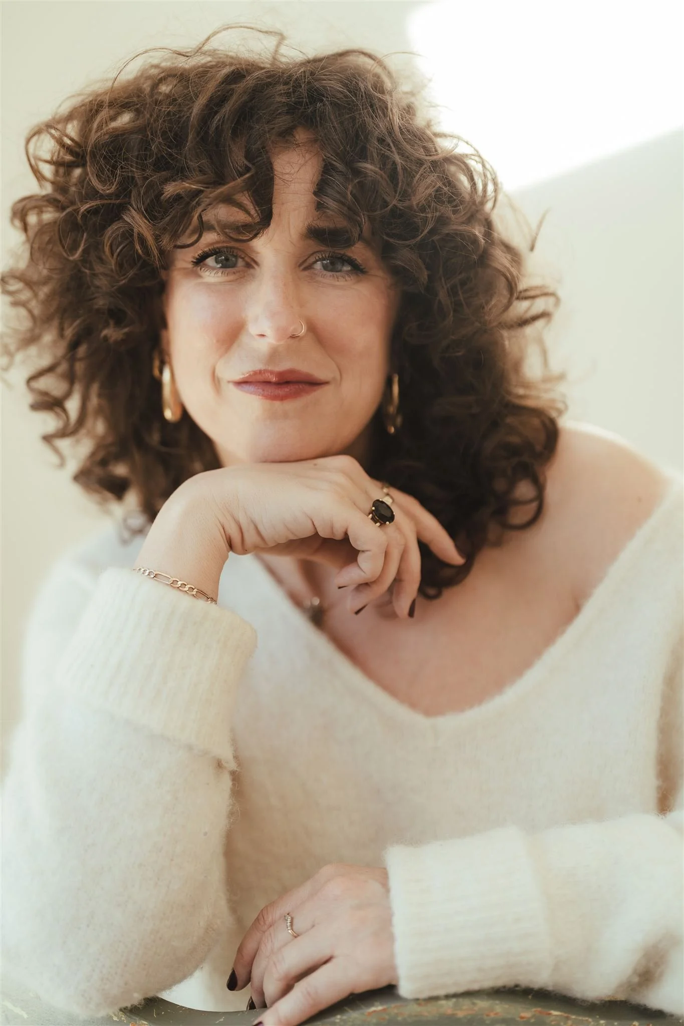 A woman with curly brown hair and fair skin rests her chin on her hand, wearing a cream-colored sweater, jewelry including rings, a bracelet, earrings, and a nose piercing, with a calm expression and direct gaze.