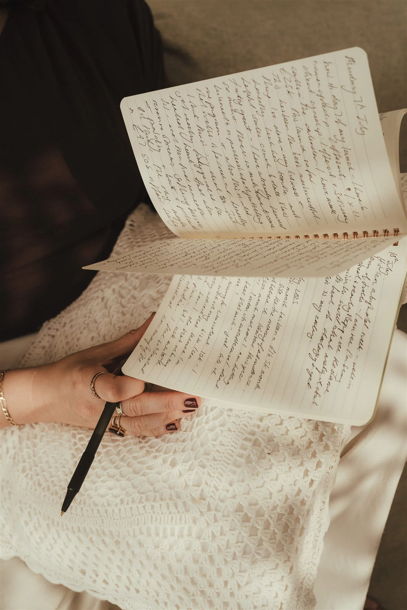 A person with dark nail polish and multiple rings on their fingers, holding a black pen in their right hand, writing in a spiral notebook that is resting on their lap. The person is wearing a cream-colored, textured knit sweater.