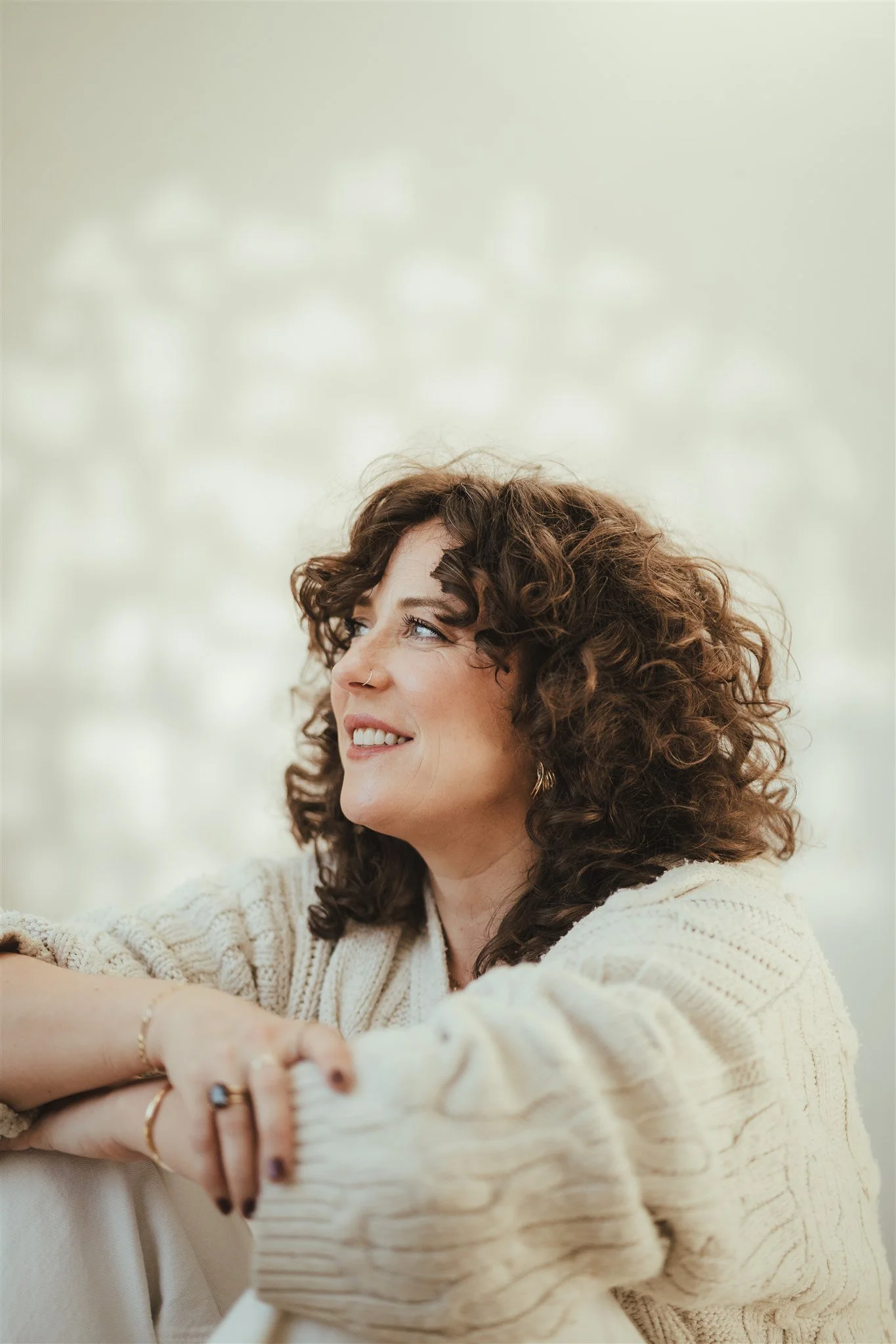 Close-up of a smiling woman with curly brown hair wearing a cream-colored sweater, sitting with her arms crossed, against a light background.