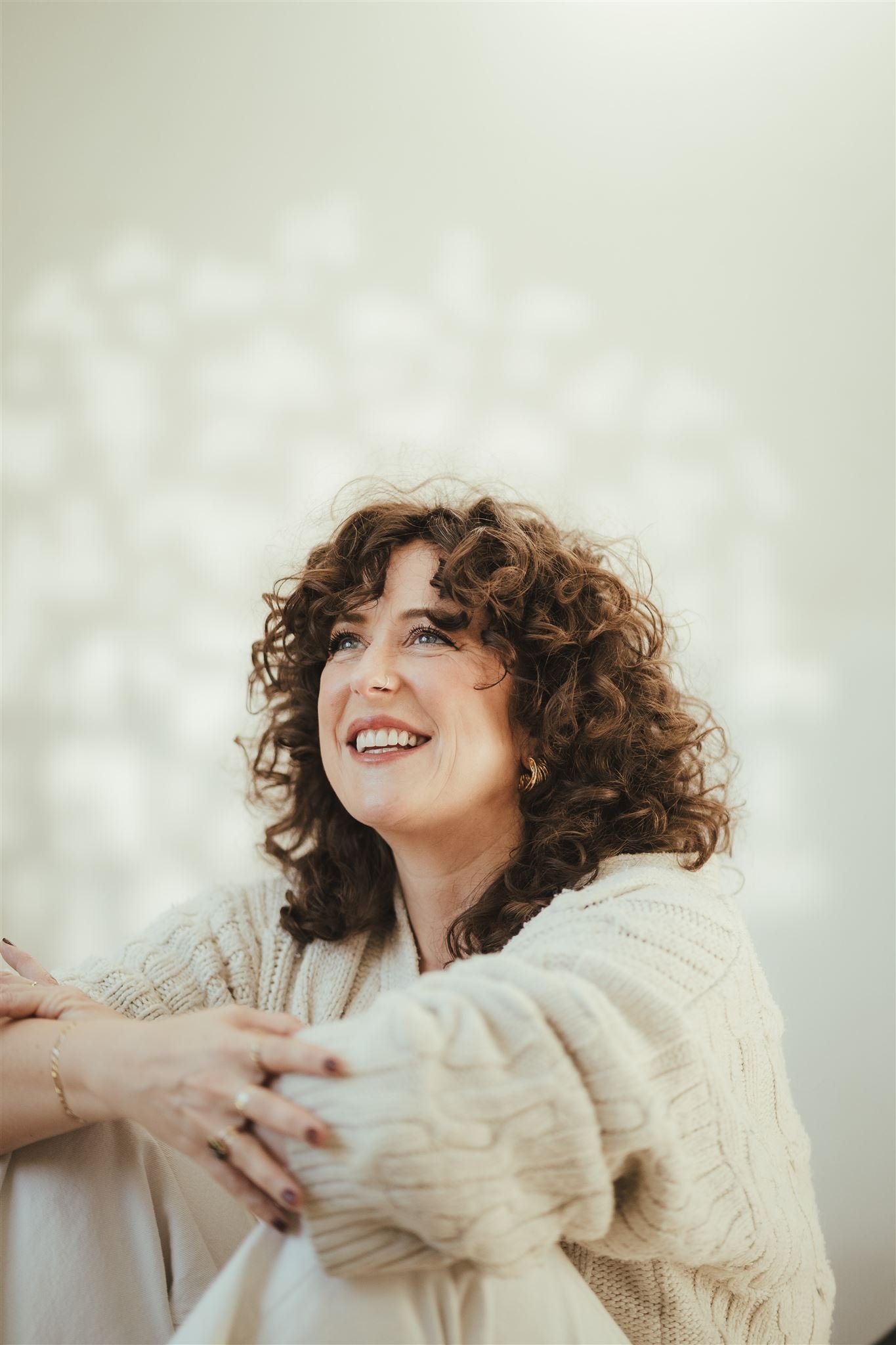 A woman with curly brown hair smiling, wearing a cream-colored sweater, sitting cross-legged outside with a blurred light-colored background.