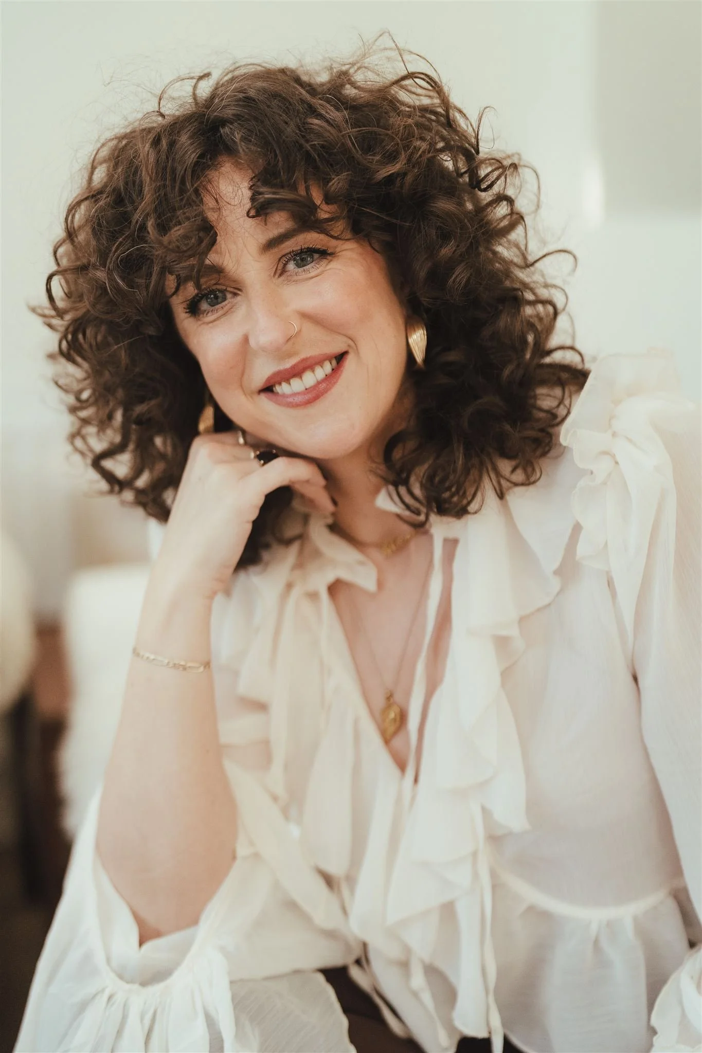 A woman with curly dark hair, smiling, wearing beige clothing with ruffled details, gold earrings, and a nose ring, sitting indoors.