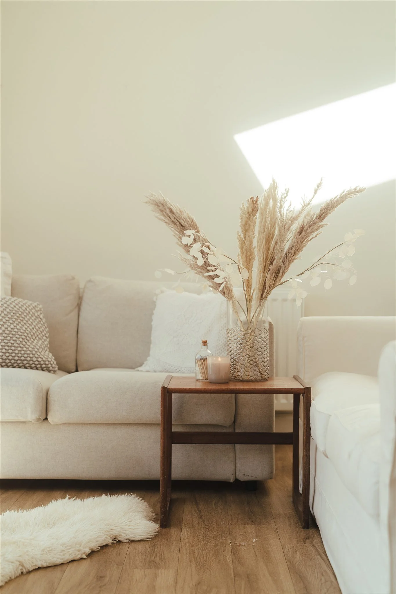 A living room with a beige sofa adorned with textured and plain throw pillows, a small wooden side table with a vase of dried pampas grass, a candle, and decorative leaves. The floor is wooden, and there's a white fluffy rug. The wall is light-colored, with sunlight streaming through a window, creating a bright atmosphere.