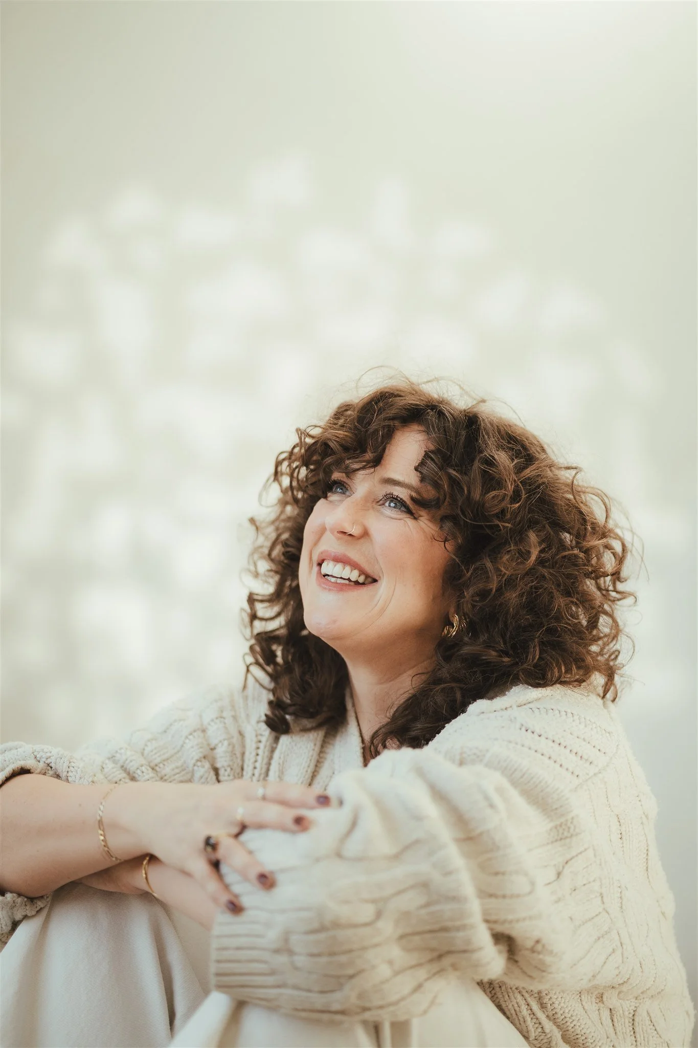A woman with curly hair and a warm smile sitting outdoors, wearing a cream-colored sweater, with a blurred natural background.