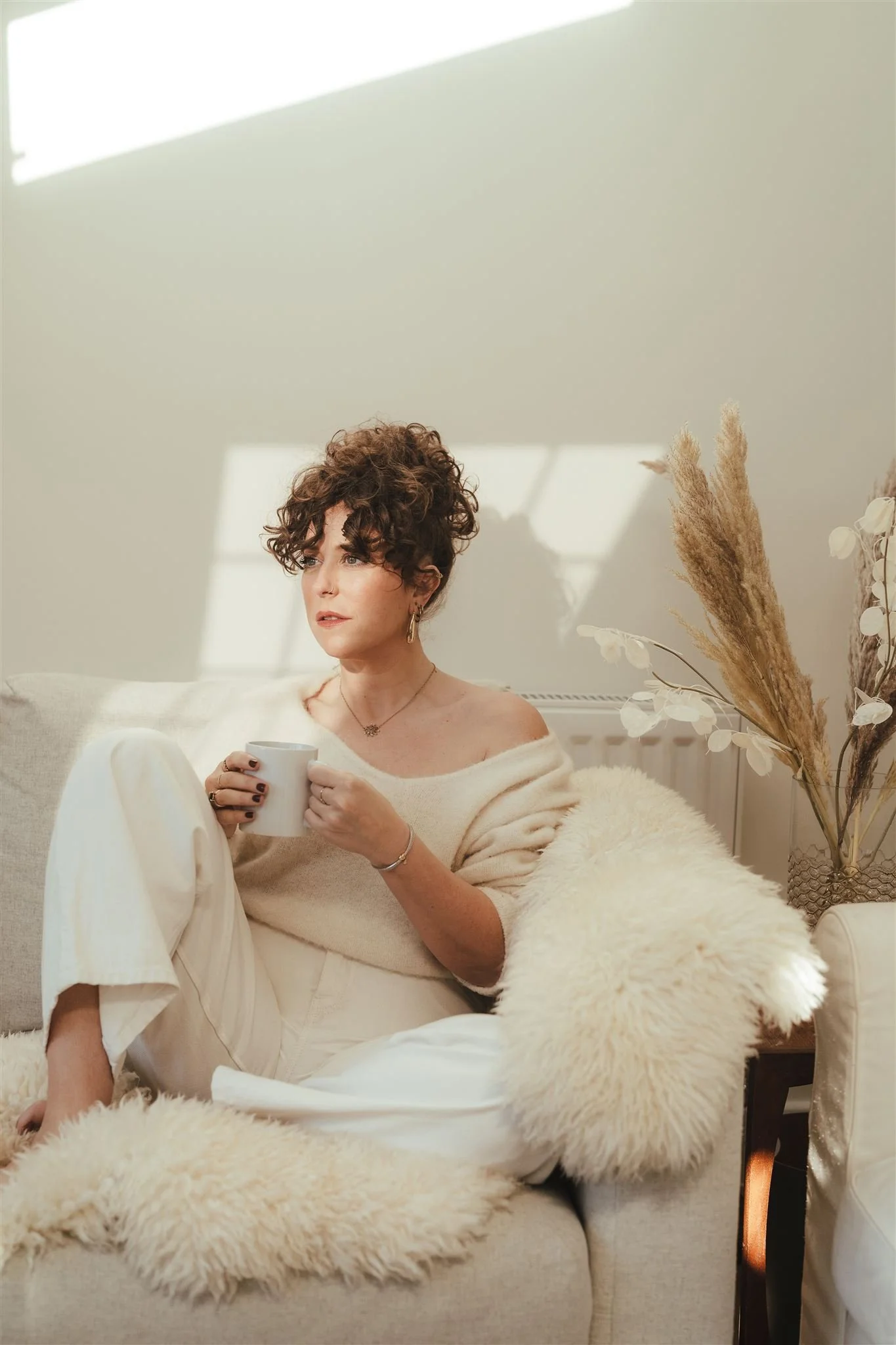 A woman with short curly brown hair sitting on a beige sofa, holding a white mug, wearing a cozy cream-colored sweater and matching pants, surrounded by a cream-colored faux fur blanket, with dried pampas grass and white flowers in a clear vase nearby, sunlight streaming through a window casting shadows.
