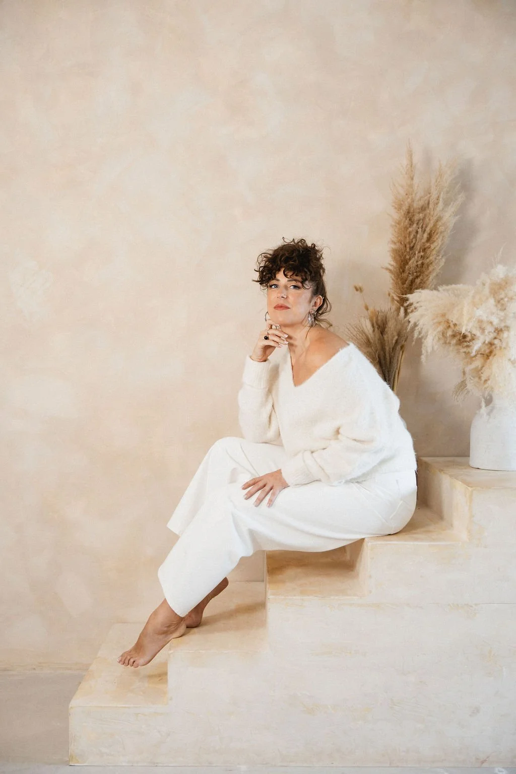 A woman with curly hair sitting on stone steps in a minimalistic indoor setting with a beige background and pampas grass in a white vase.