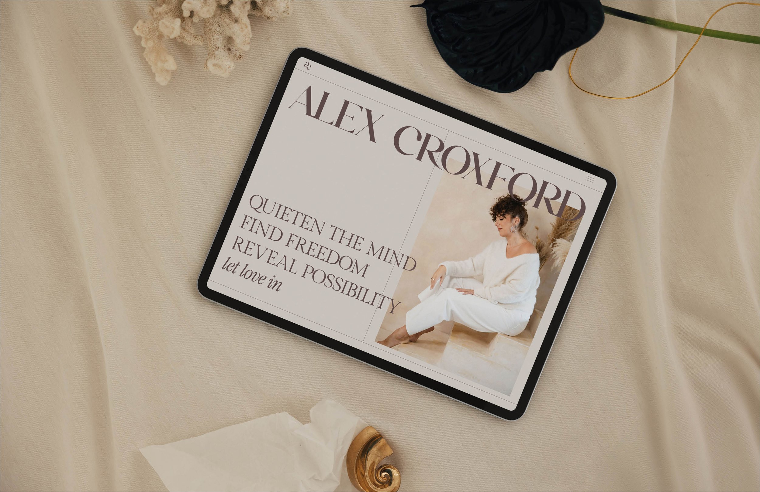 A tablet displaying an advertisement for Alex Croxford, featuring a woman in white seated on a light wooden chair, with text emphasizing quiet and freedom, placed on a beige fabric surface along with decorative items like a shell, black fabric, a dried plant, and a spiral ornament.