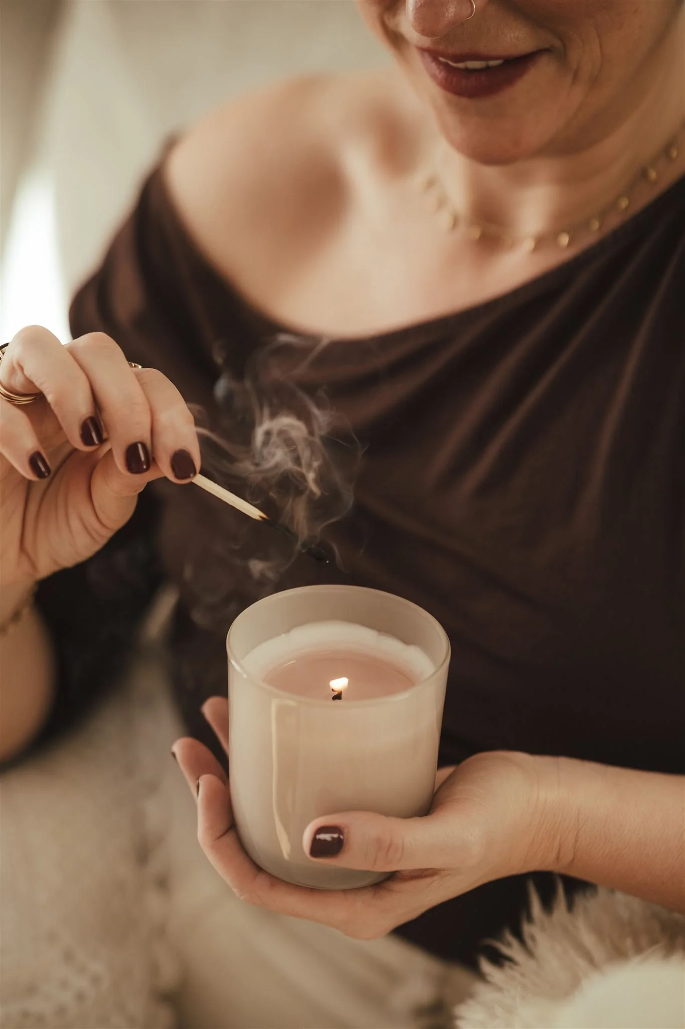A woman with dark nail polish holding a lit candle and a matchstick near her face, with visible smoke. She is wearing a black top and a gold necklace, and the background includes a fuzzy surface and part of a dog.