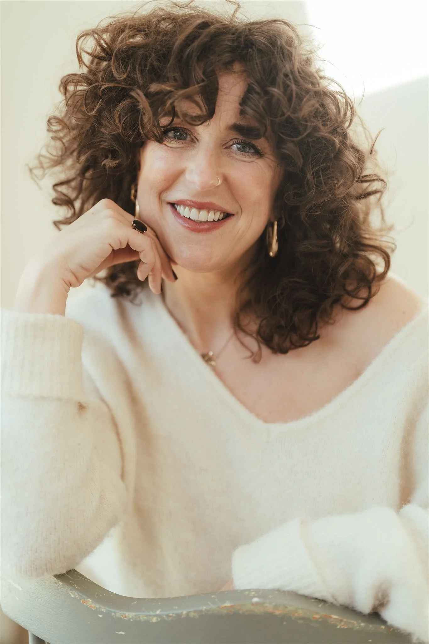 A woman with curly brown hair smiling, wearing earrings and a nose ring, resting her chin on her hand, dressed in a white sweater.