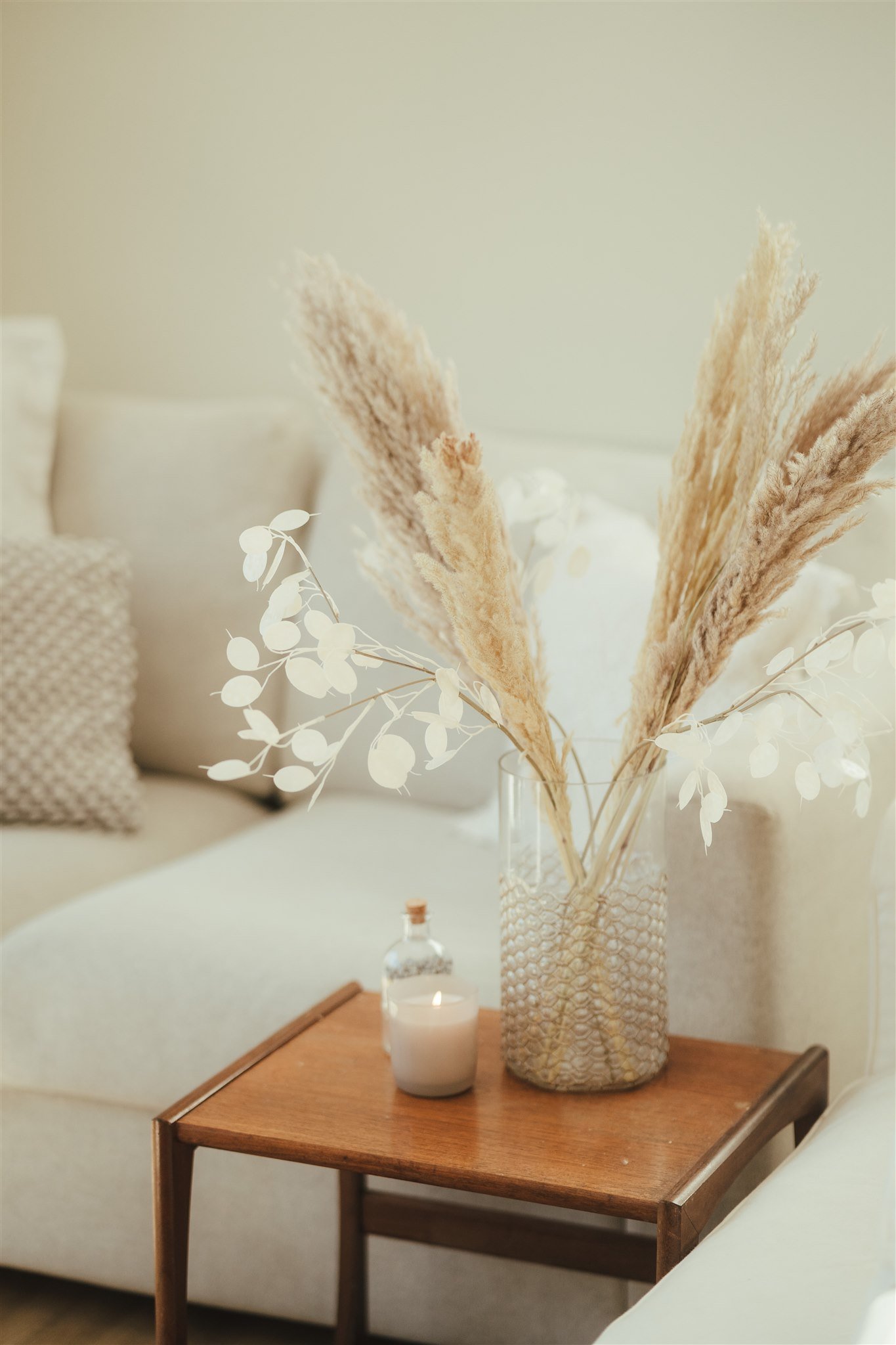 A wooden side table with a tall textured vase containing dried pampas grass and white leaves, a white candle in a glass jar with a cork lid, set against a neutral-colored background with a sofa and pillows.