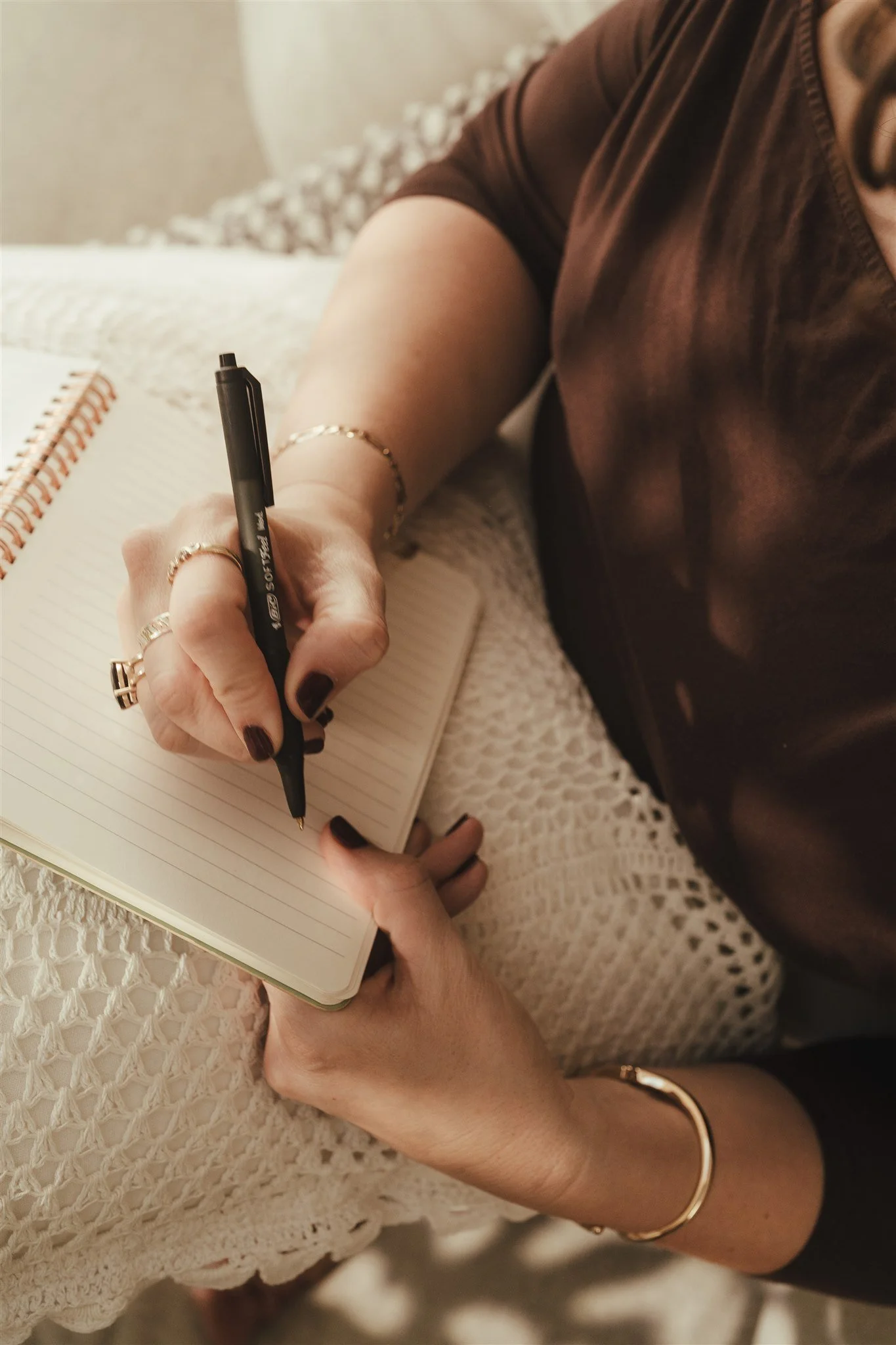 A person writing in a notebook with a pen, wearing jewelry such as rings, a bracelet, and a bangle, sitting on a crocheted blanket.