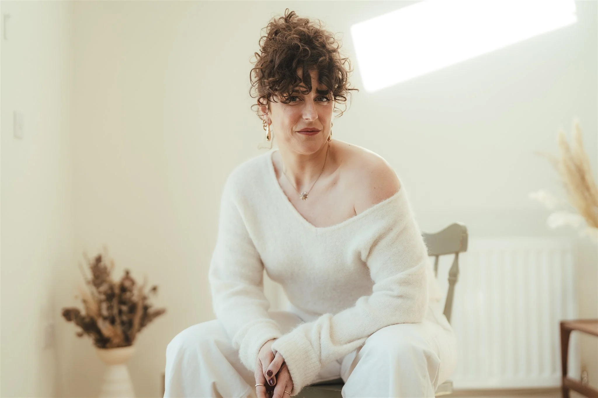 A woman with curly brown hair wearing a white off-shoulder sweater and matching pants, sitting on a chair in a bright room with minimal decor and a dried plant in the background.