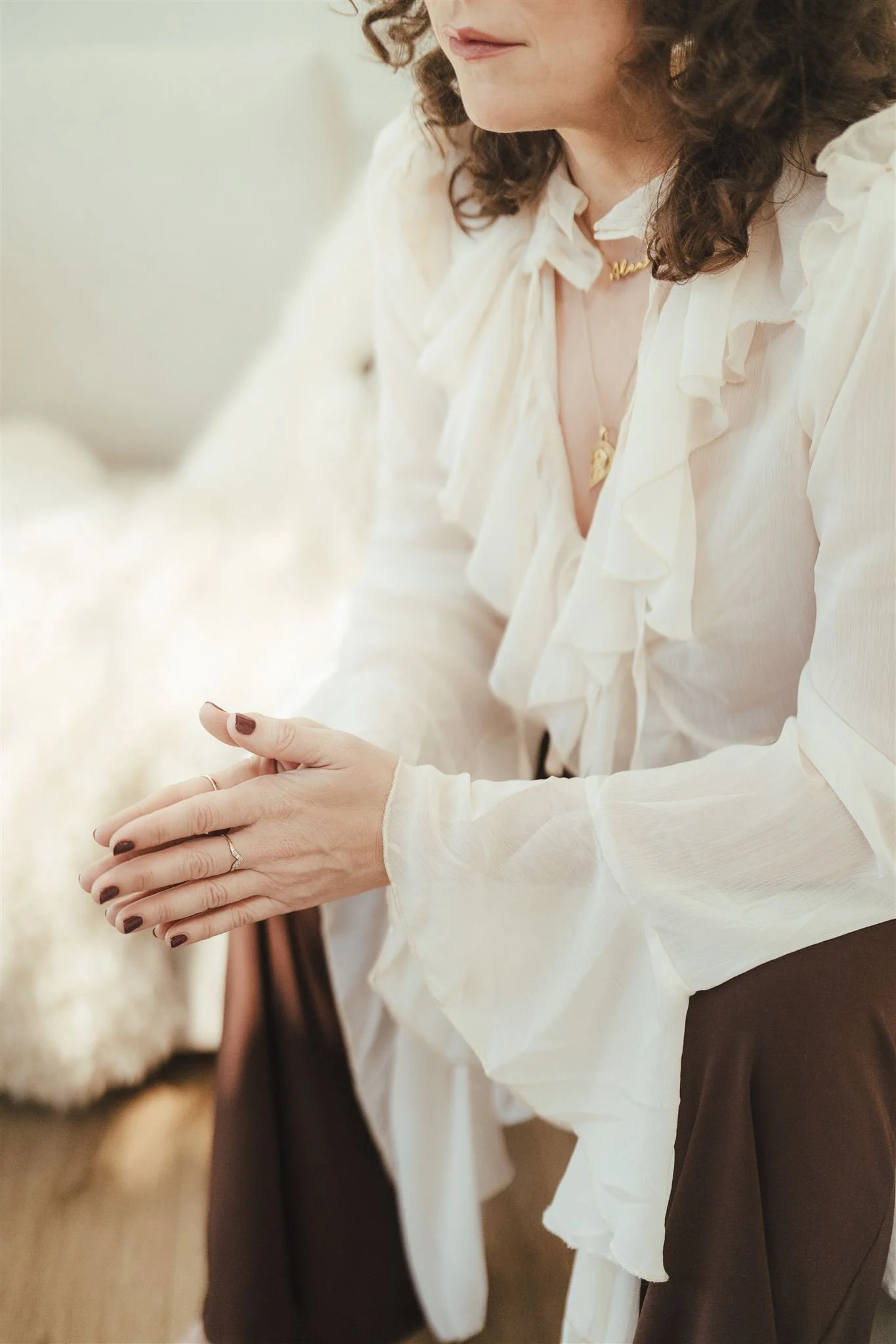 Close-up of a woman with curly hair wearing a white blouse with ruffles, a gold necklace, and a ring, holding a smartphone.
