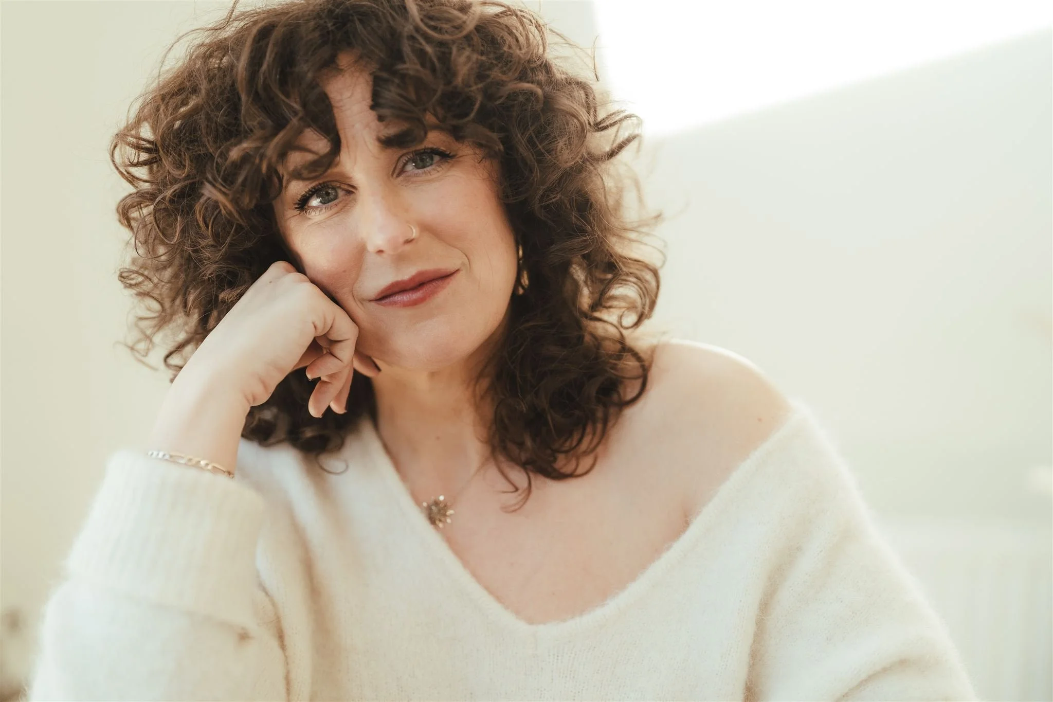 A woman with curly brown hair and hazel eyes resting her chin on her hand, wearing a white sweater with a gold bracelet and necklace, in front of a light background.