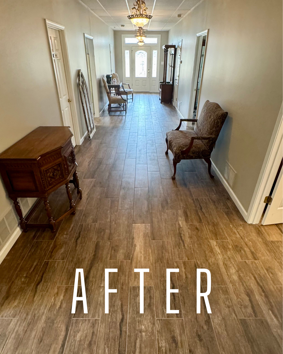 A hallway with wood-like tile flooring, beige walls, and antique-style furniture including chairs and a small table. The area is well-lit with decorative ceiling lights, and there is a front door with glass panels at the end.