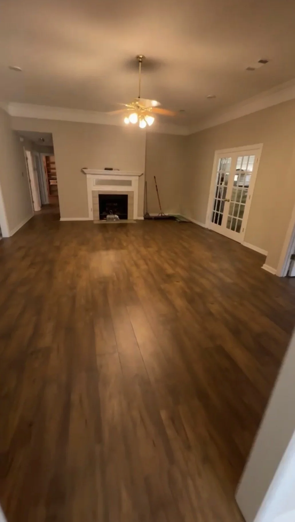 Empty living room with hardwood floors, a ceiling fan with lights, a white fireplace, and glass double doors leading outside.