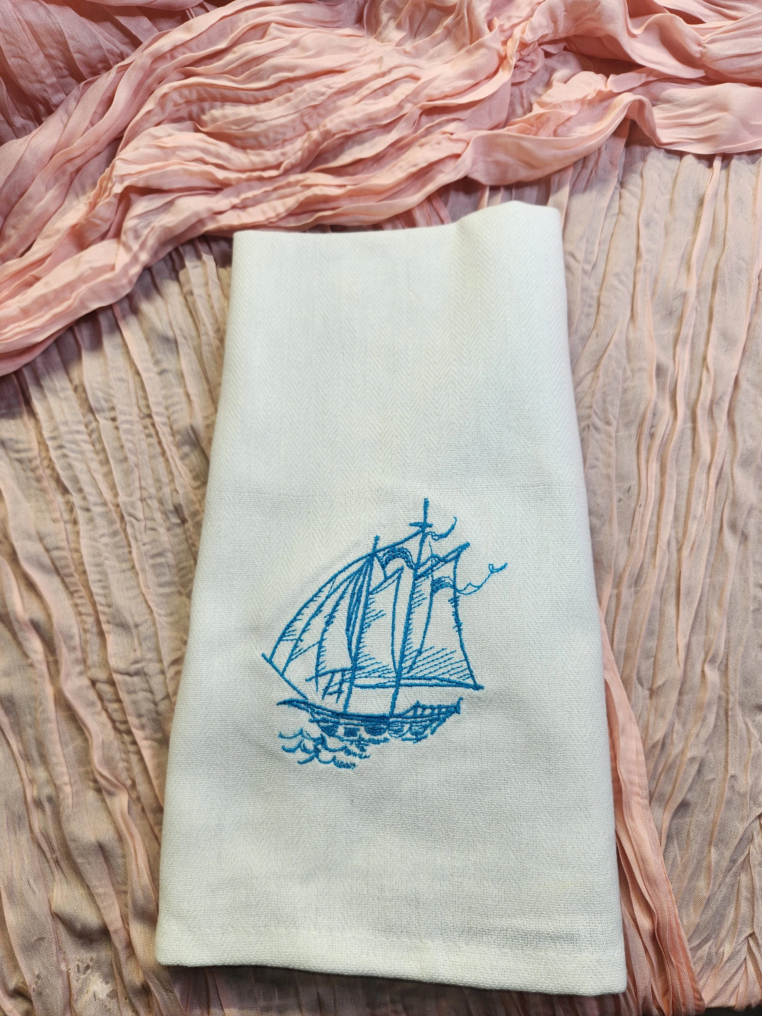 A white cloth with a blue embroidered sailboat design, placed on a bed with pink and beige wrinkled sheets.