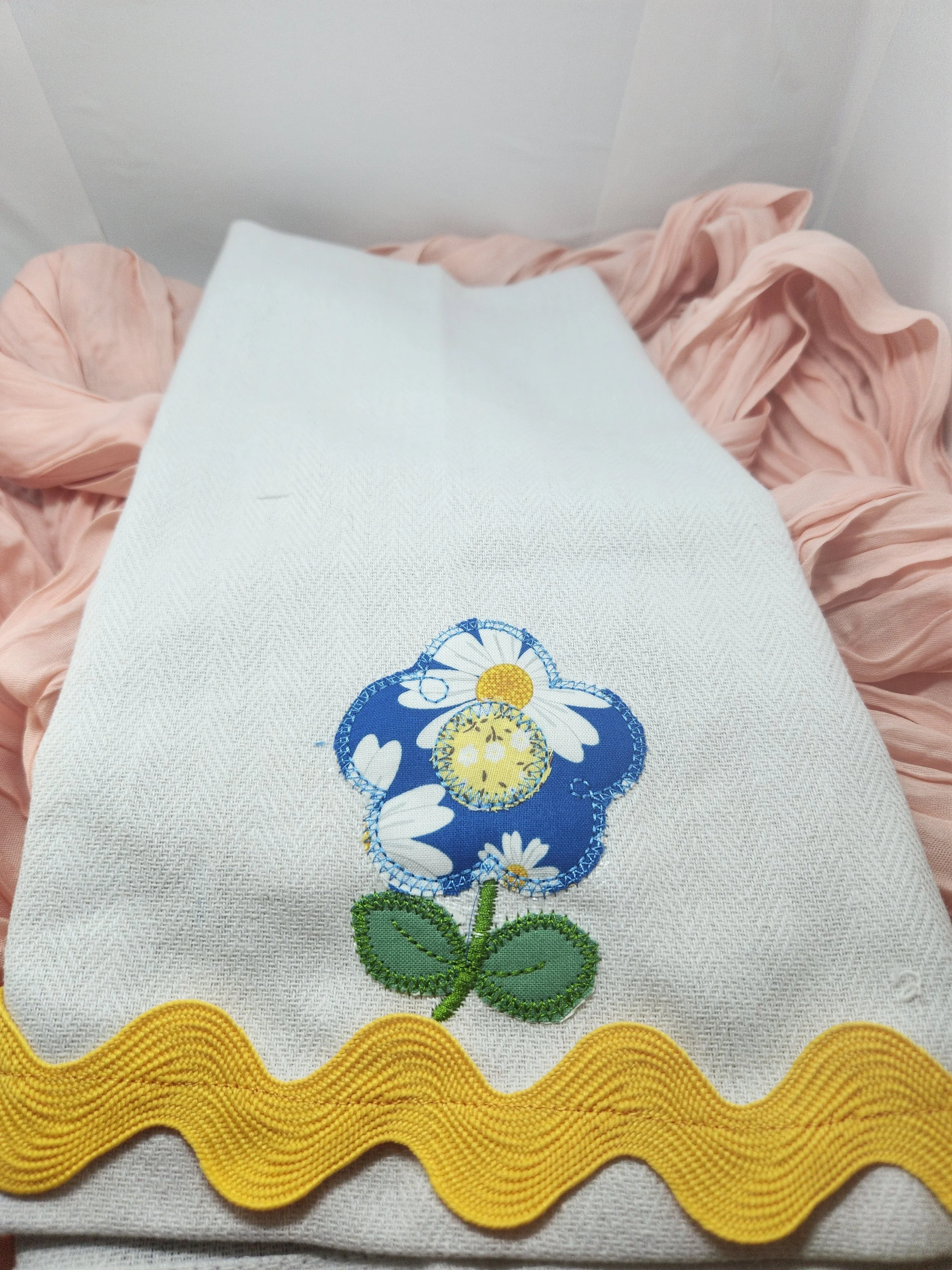 A white fabric item with a colorful embroidered flower featuring blue petals with white daisies, green leaves, and yellow accents, bordered at the bottom with yellow wavy trim, placed on pink ruffled fabric.