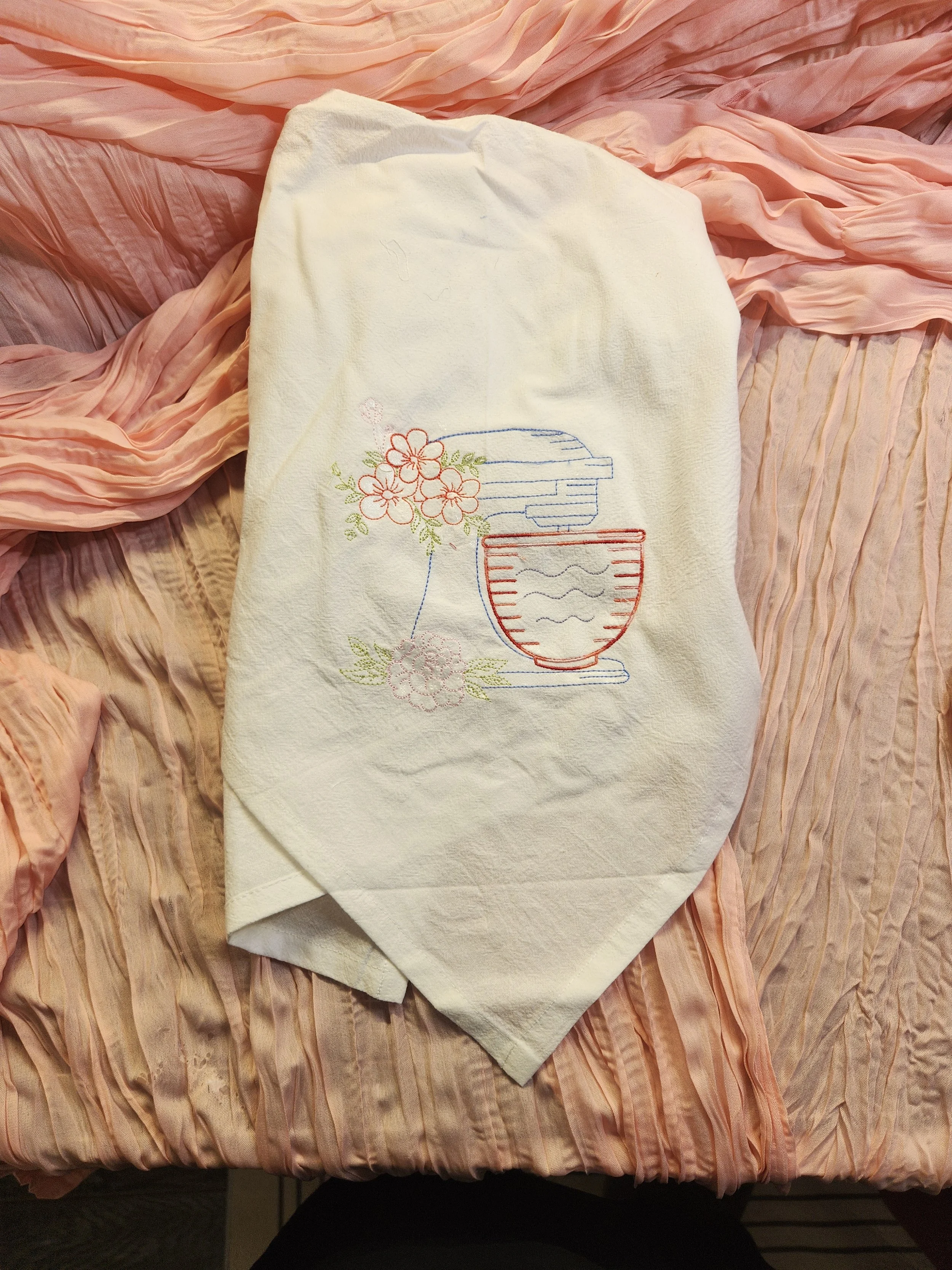Embroidered dish towel with a mixer and flowers, placed on a pink bedspread.