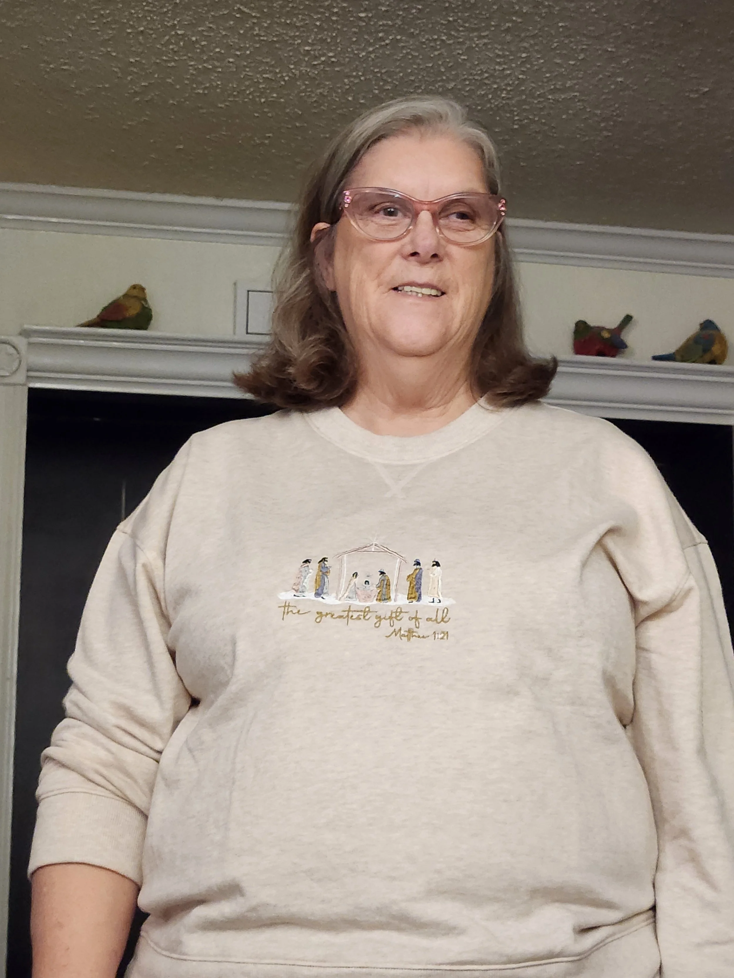 A middle-aged woman with gray hair, wearing glasses and a cream sweatshirt with an embroidered nativity scene and the quote 'the greatest gift of all' from Matthew 2:10. She is standing indoors in front of a black fireplace with decorative bird figurines on the mantel.