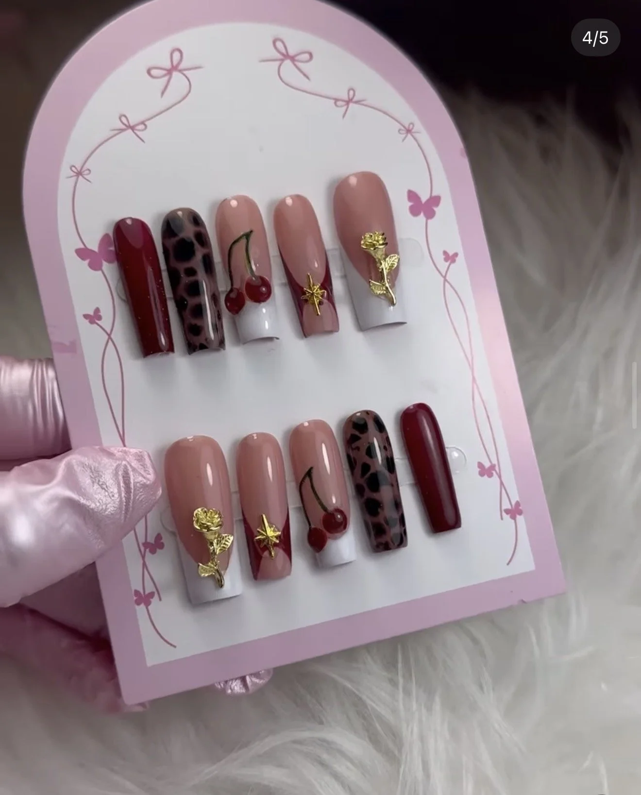 Set di unghie finte con vari design, tra cui righe, leopardato, ciliegie, e decorazioni dorate, su una confezione rosa con motivo di farfalle.