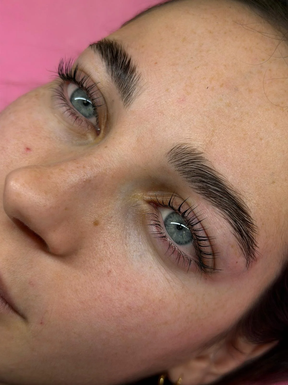 Primo piano di un volto di una donna con occhi azzurri, sopracciglia folte e ciglia lunghe, con uno sfondo rosa.