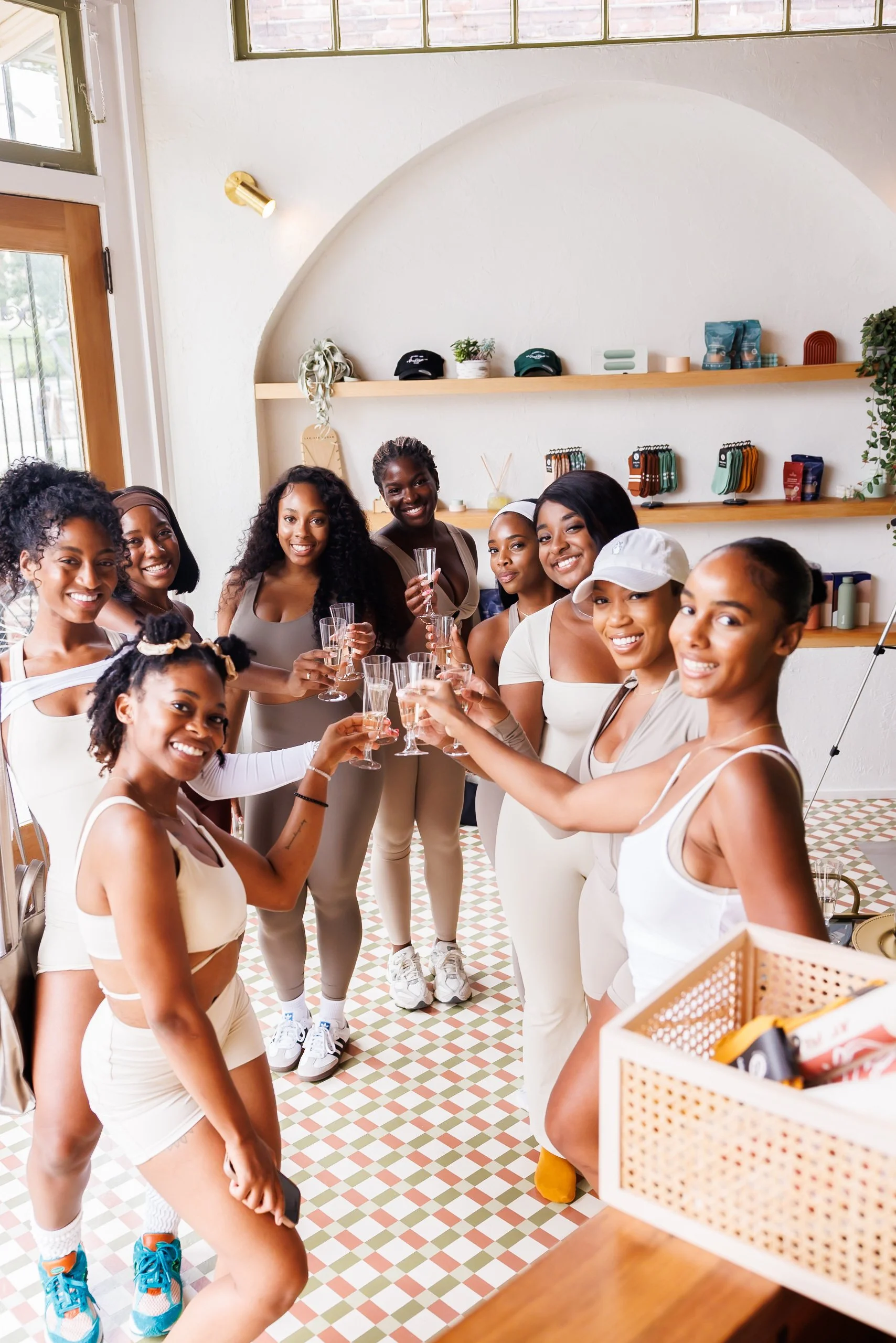 A group of women in casual workout clothes toasting with champagne glasses inside a brightly lit room with white walls, wooden shelves, and a patterned tile floor.