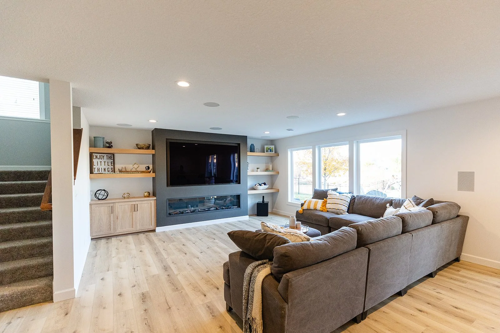 Living room with a large sectional sofa, a wall-mounted TV, a modern fireplace, shelves with decorative items, large windows, and wooden flooring.