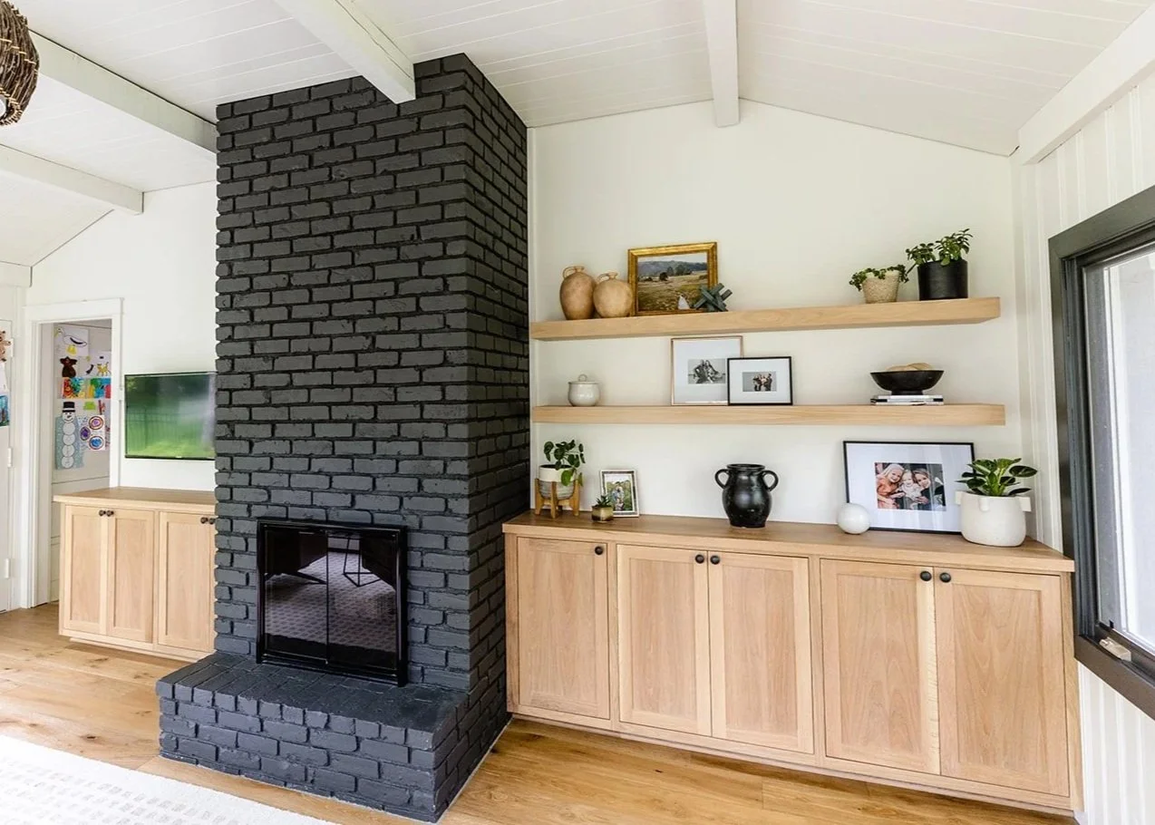 Living room with a black brick fireplace, wooden cabinets, and open shelves decorated with framed photos, potted plants, vases, and other decor.