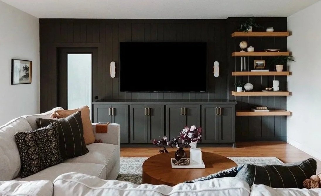 Living room with white sofa, dark accent wall with mounted flat-screen TV, wooden floating shelves with decorations, round wooden coffee table with flower vase, hardwood floor, and framed wall art.