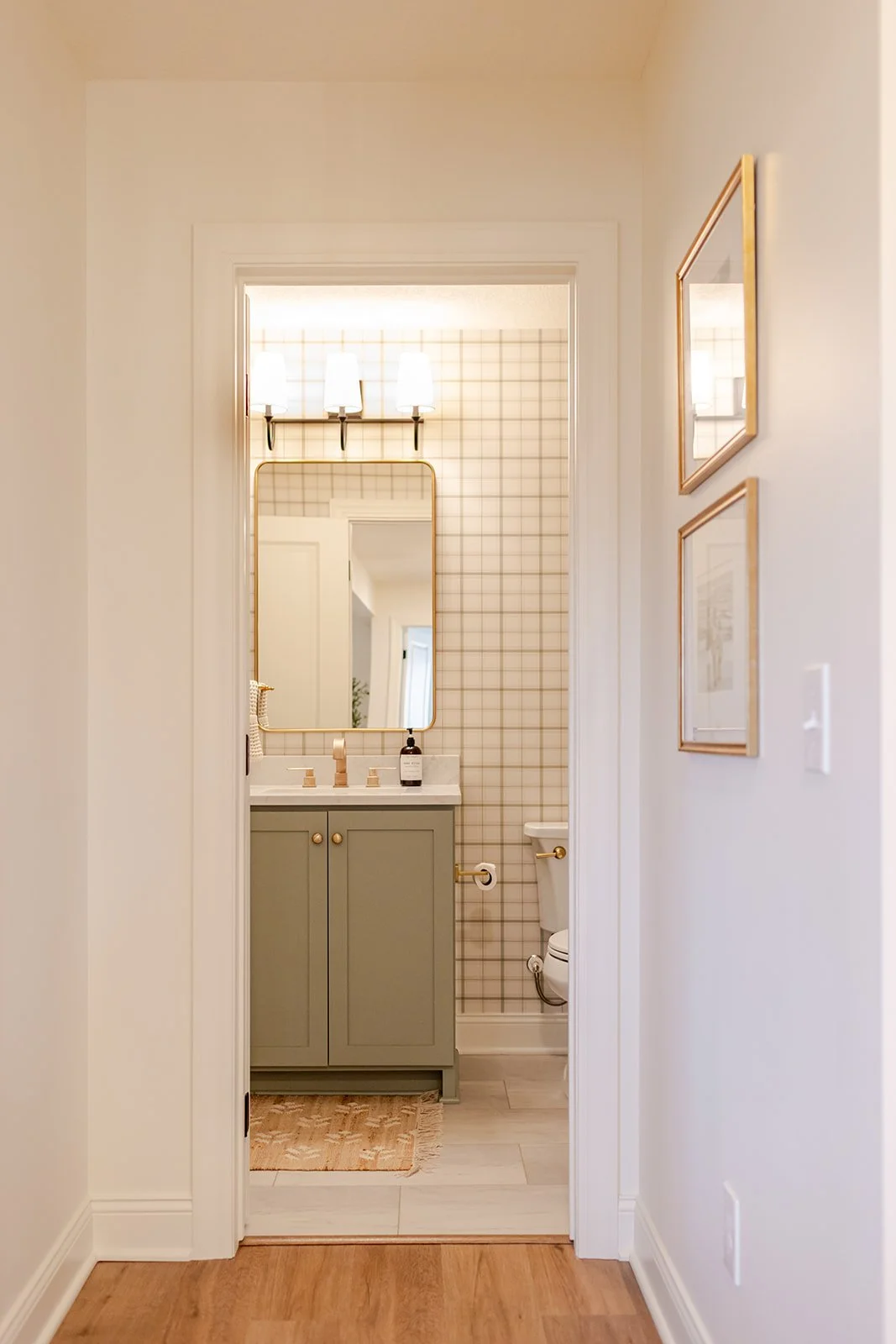 View of a small bathroom from a hallway, featuring a green vanity with a white countertop, a mounted mirror, a wall-mounted light fixture, a framed picture on the wall, and a partial view of the toilet.
