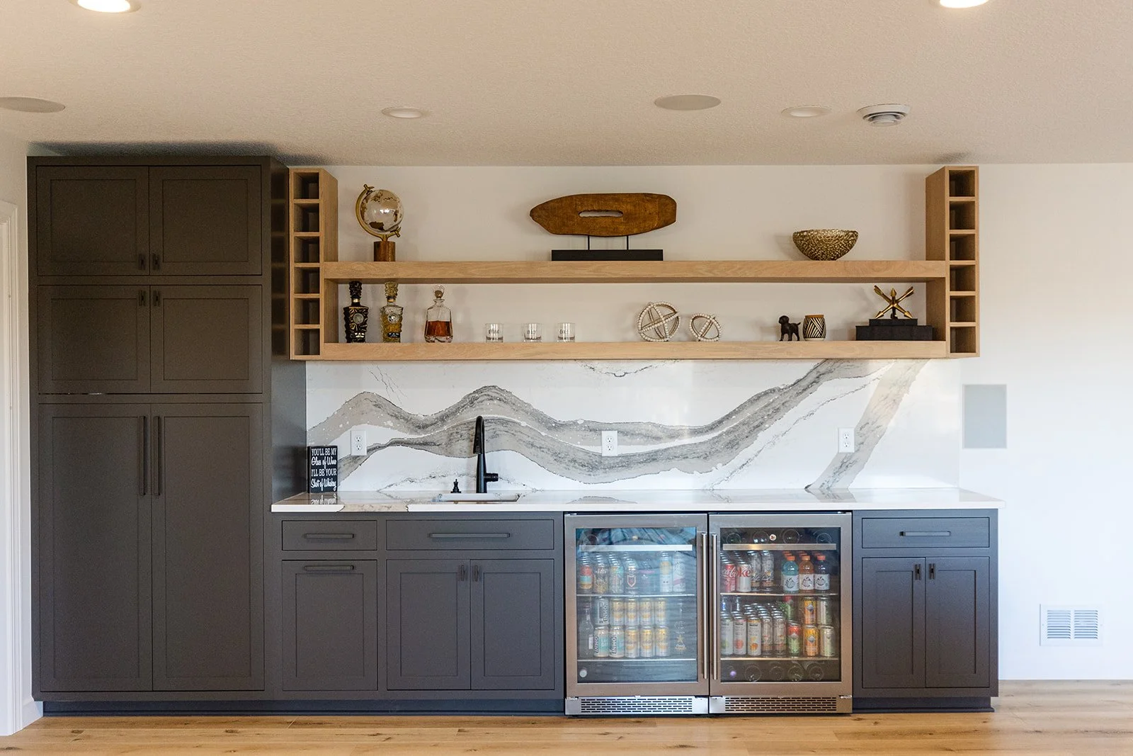 Modern kitchen with gray cabinets, marble backsplash, open wooden shelves, and a mini refrigerator filled with drinks.