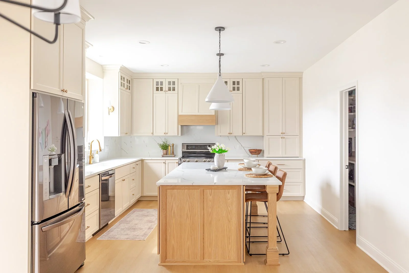 Bright kitchen with white cabinets, stainless steel refrigerator, black stove, white marble countertops, a wooden kitchen island, and four brown barstools, decorated with a white vase of green flowers and tableware.