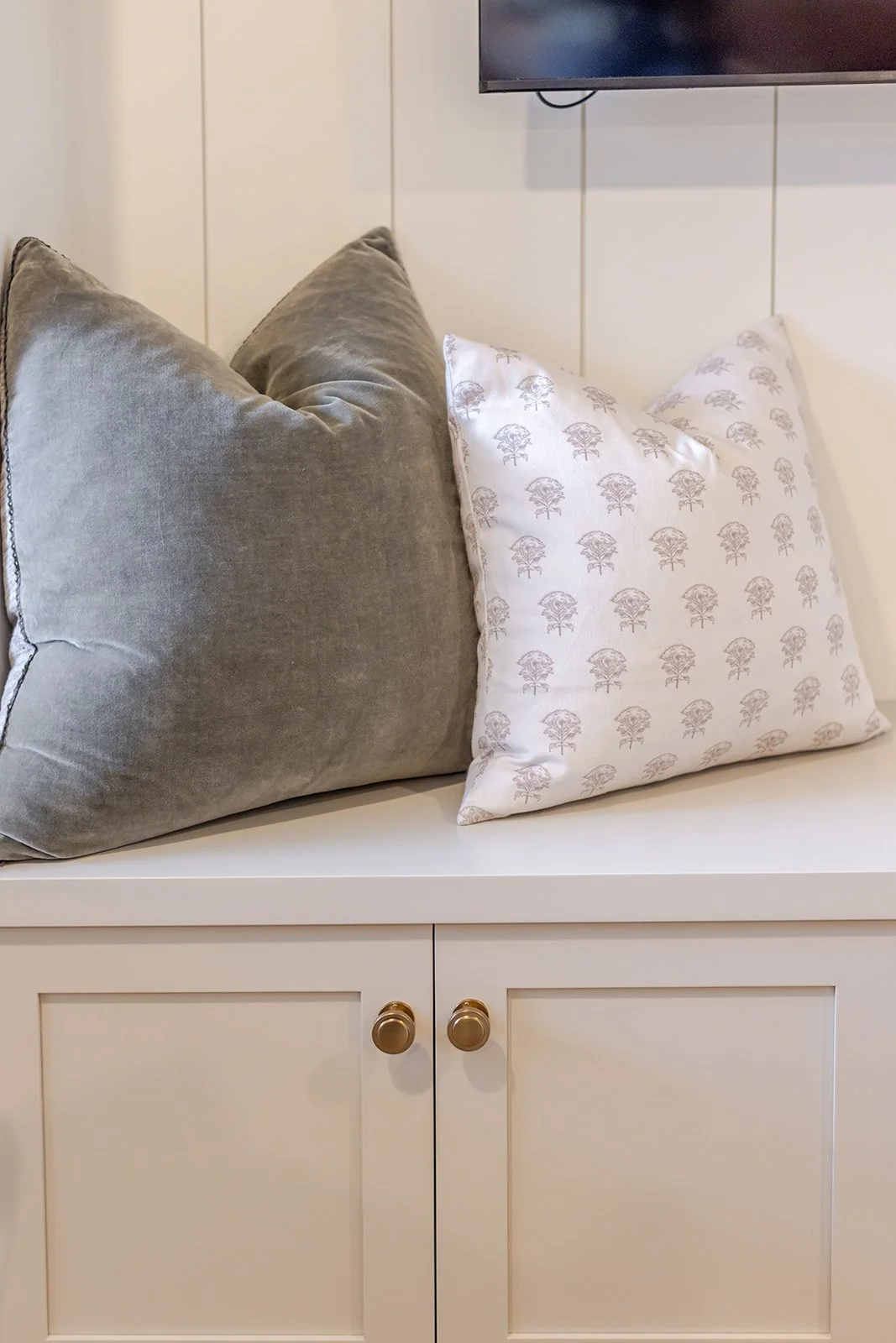 Close-up of a white cabinet with two gold knobs, topped with two pillows, one gray velvet and one white with a delicate tree pattern, in front of a wall-mounted TV.