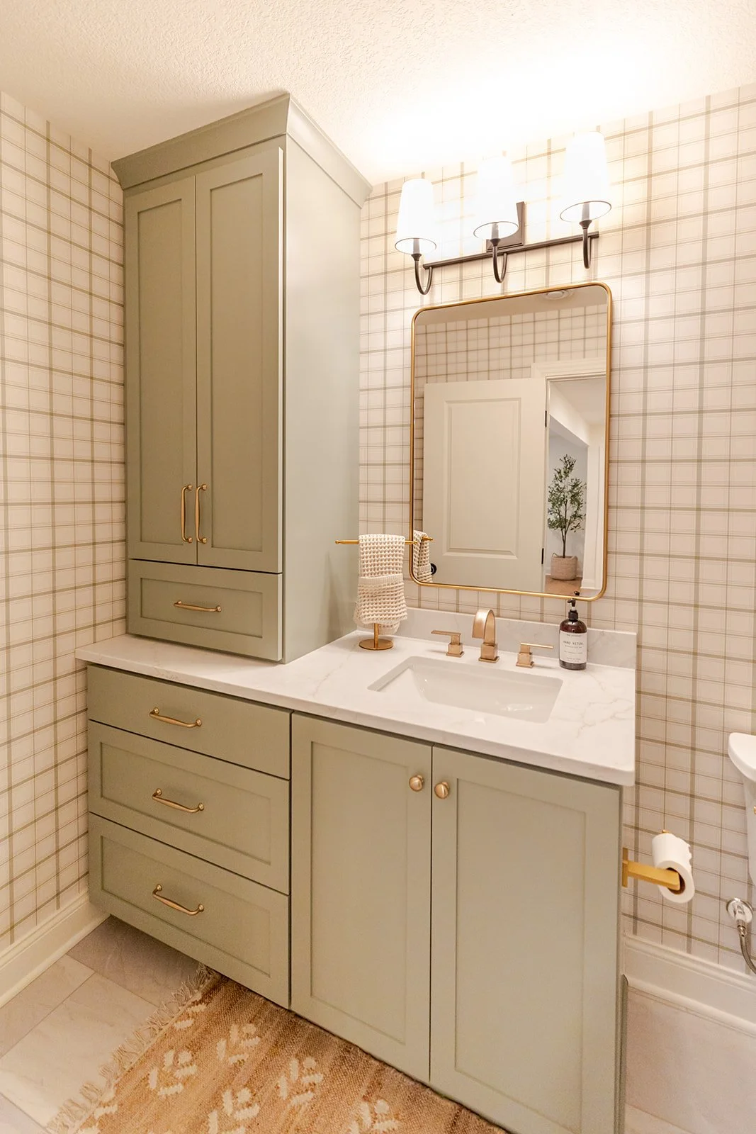 Bathroom vanity with sage green cabinets, marble countertop, gold hardware, a mirror with gold trim, and three white light fixtures, decorated in neutral tones.