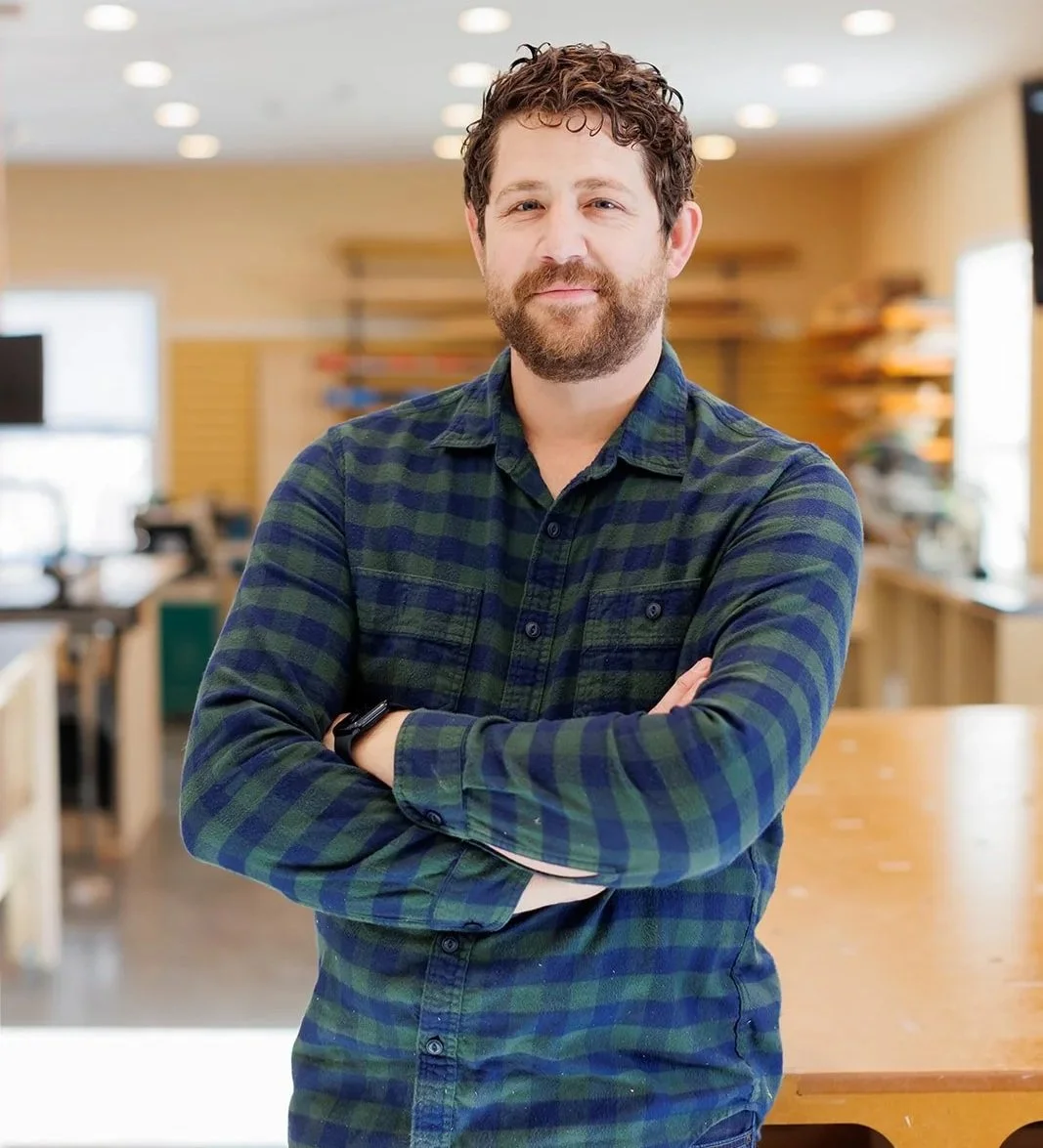 A man with a beard and curly hair standing with crossed arms in a brightly lit room, wearing a green and blue plaid shirt.