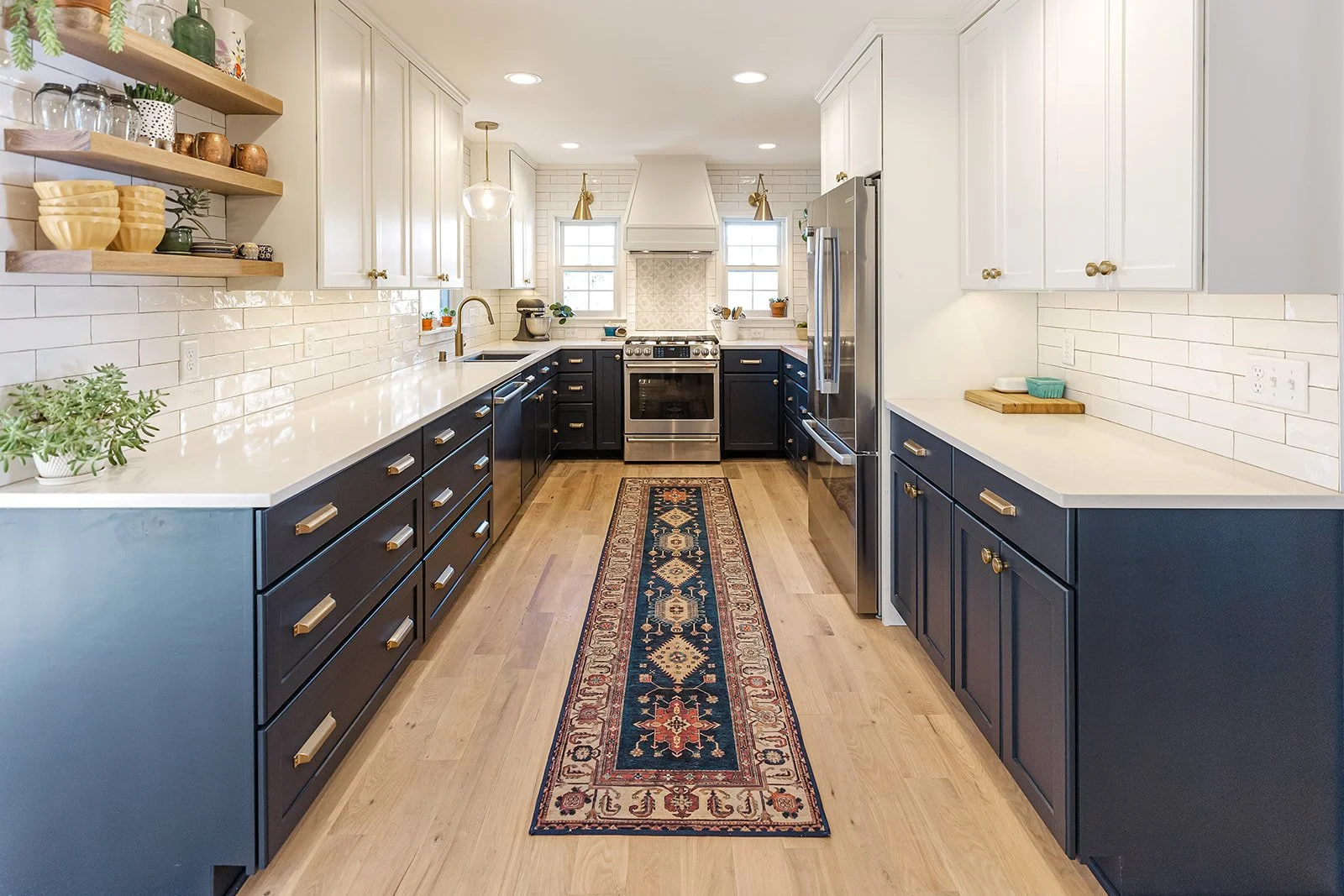 Modern kitchen with white upper cabinets, navy lower cabinets, stainless steel appliances, a long runner rug, and open wooden shelves with dishes and plants.