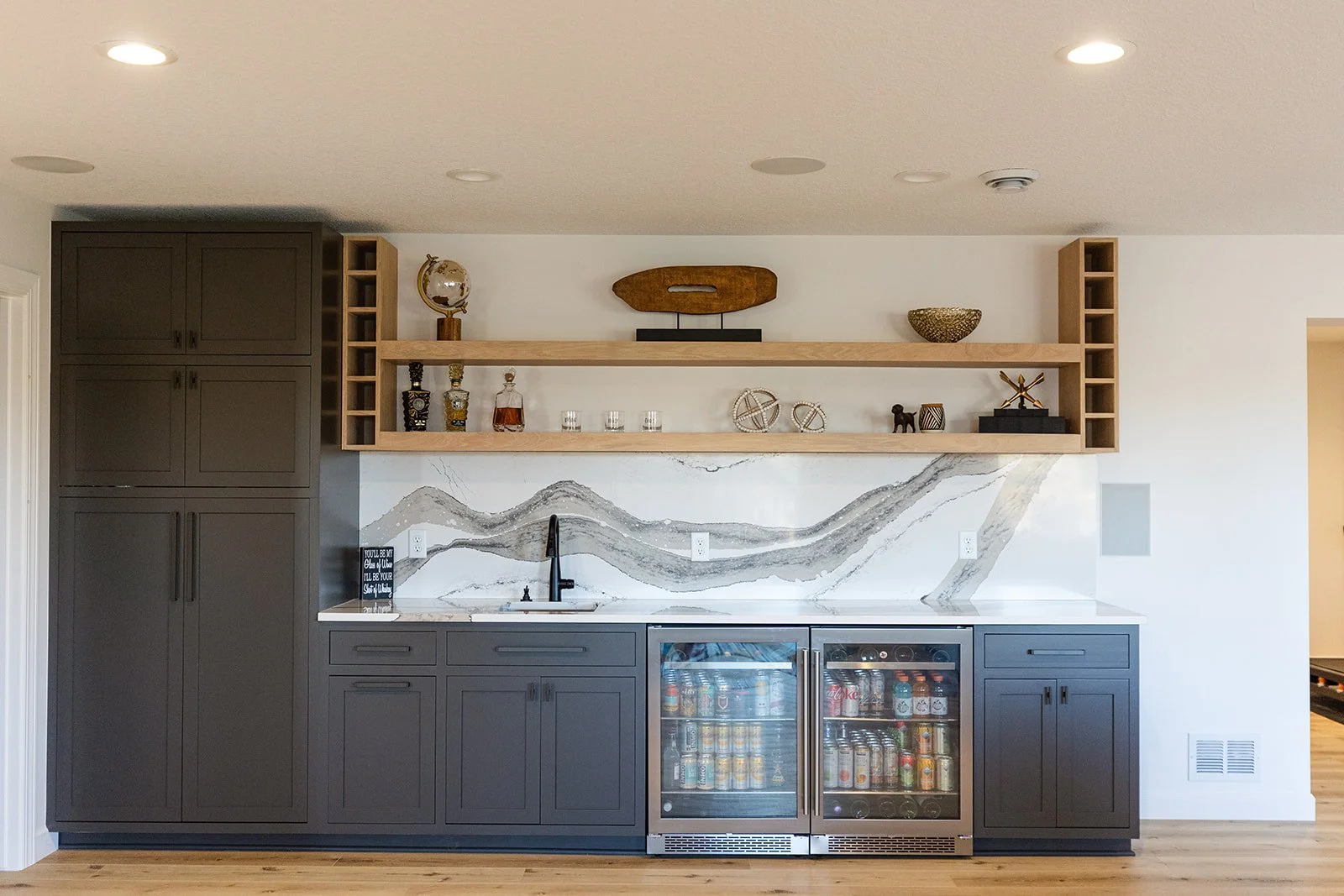 Modern kitchenette with dark gray cabinets, a white marble backsplash, open wooden shelves with decorative items, and a mini-fridge stocked with drinks.