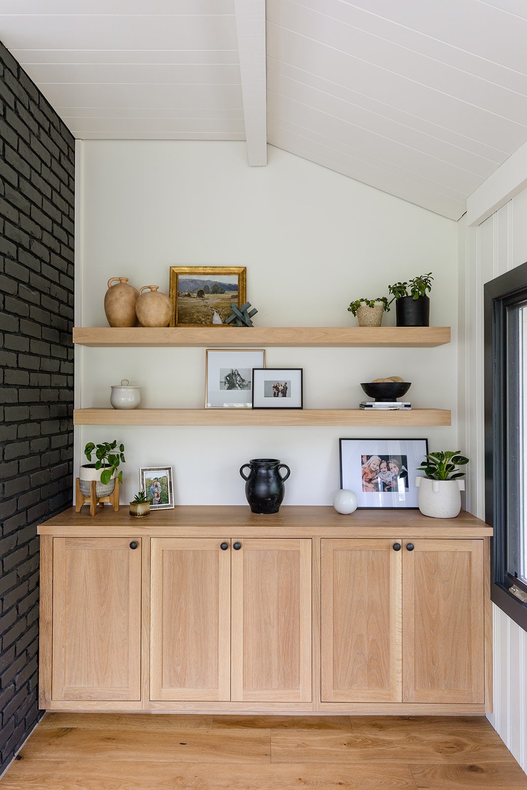 Decorative shelves with picture frames, vases, and potted plants above a wooden cabinet in a room with a black brick wall on the left and a window on the right.