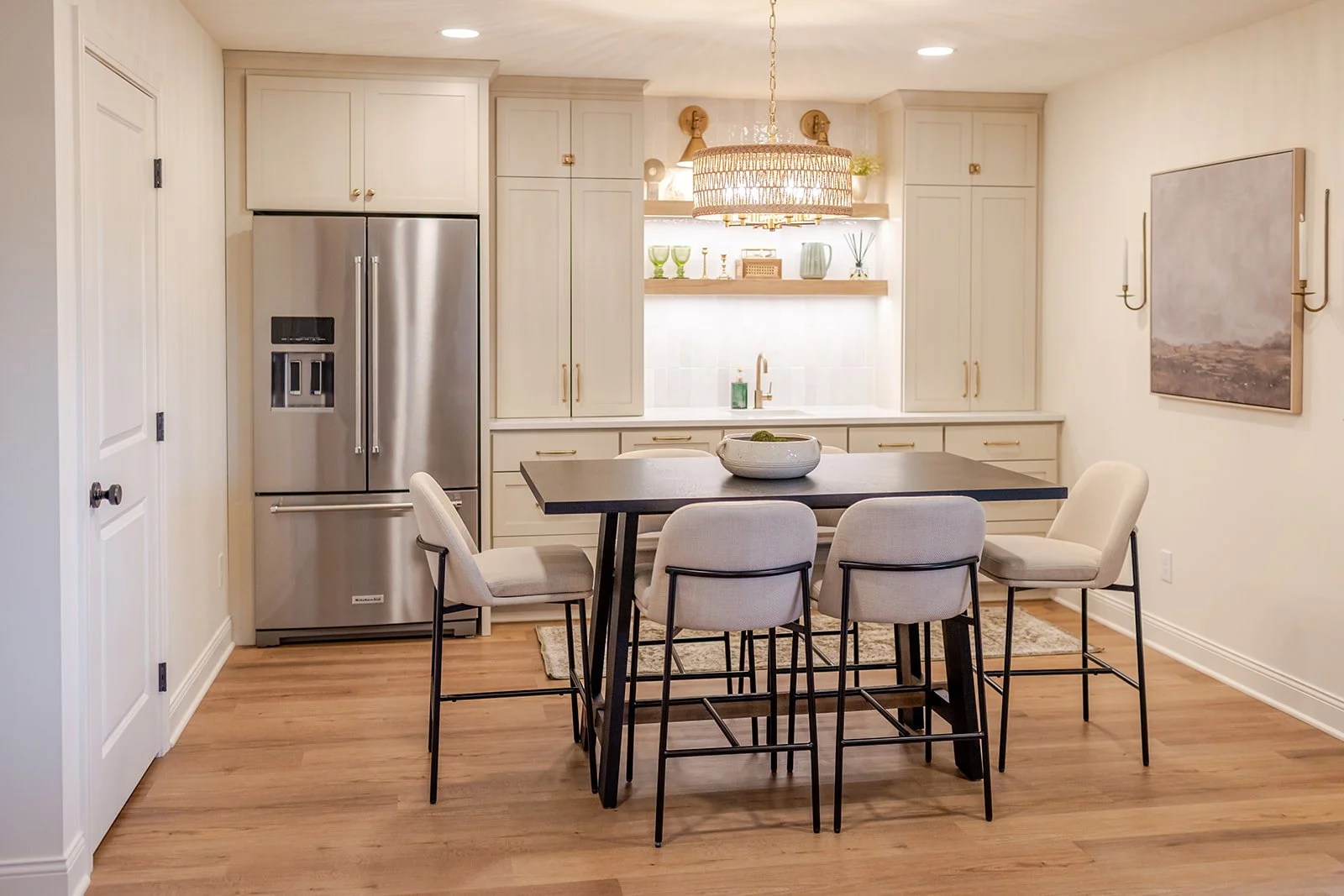 A modern kitchen with light-colored cabinetry, a stainless steel refrigerator, a dark dining table with white chairs, and decorative items on open shelves, illuminated by a large round chandelier.