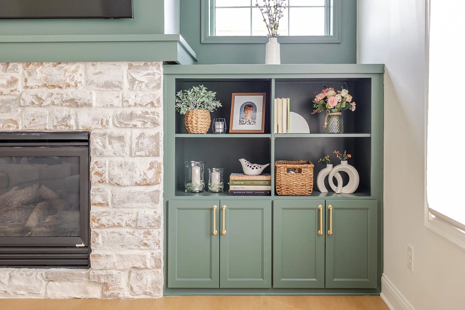 A green cabinet with decorative items and books, next to a white brick fireplace and window.