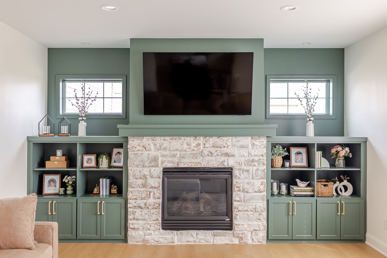 Living room with a fireplace, two green built-in shelves on either side, a large flat-screen TV above the fireplace, and decorative items including framed photos, vases with flowers, and books.