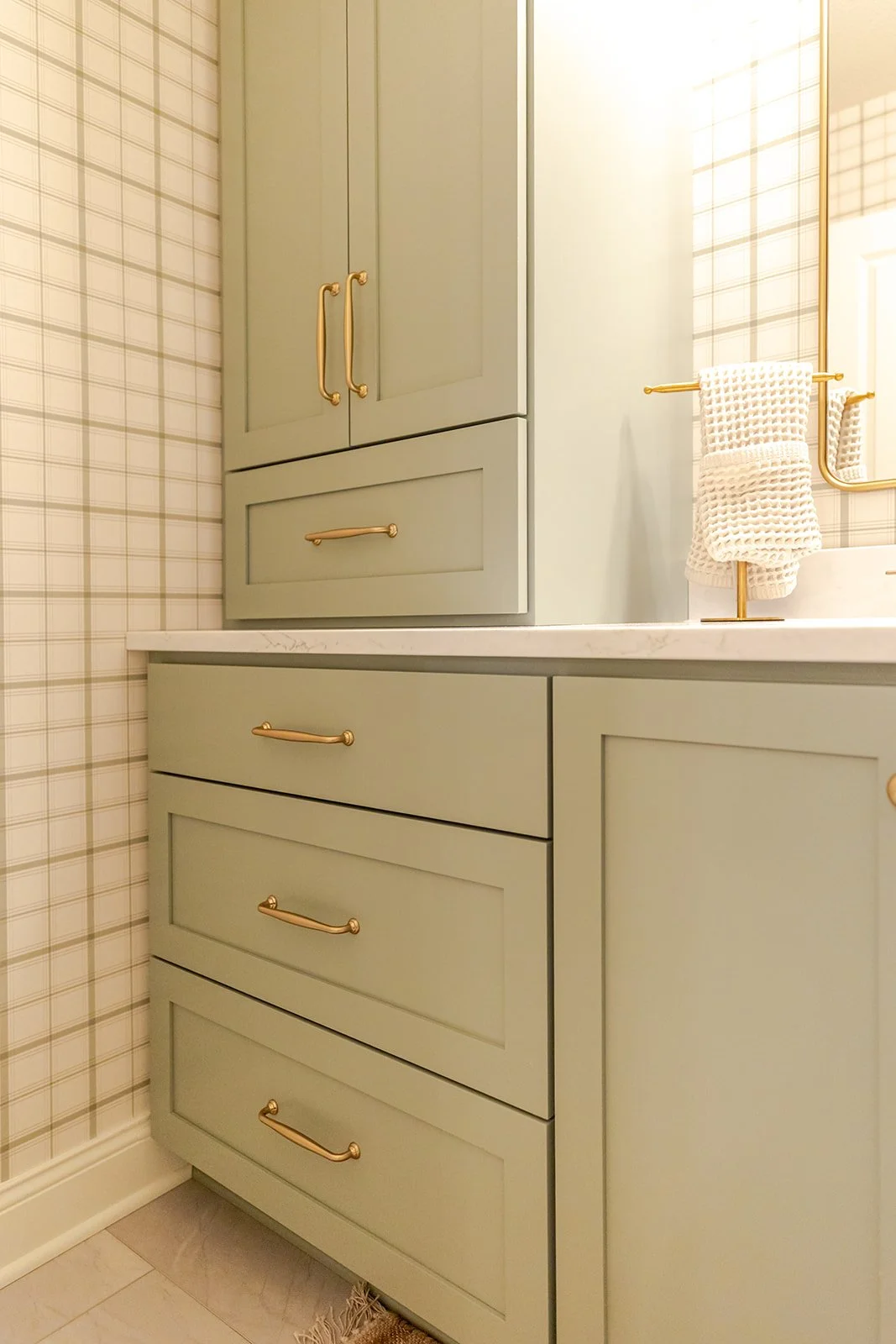 A bathroom vanity with mint green cabinetry, gold handles, and a white marble countertop, with a plaid wallpaper on the adjacent wall.
